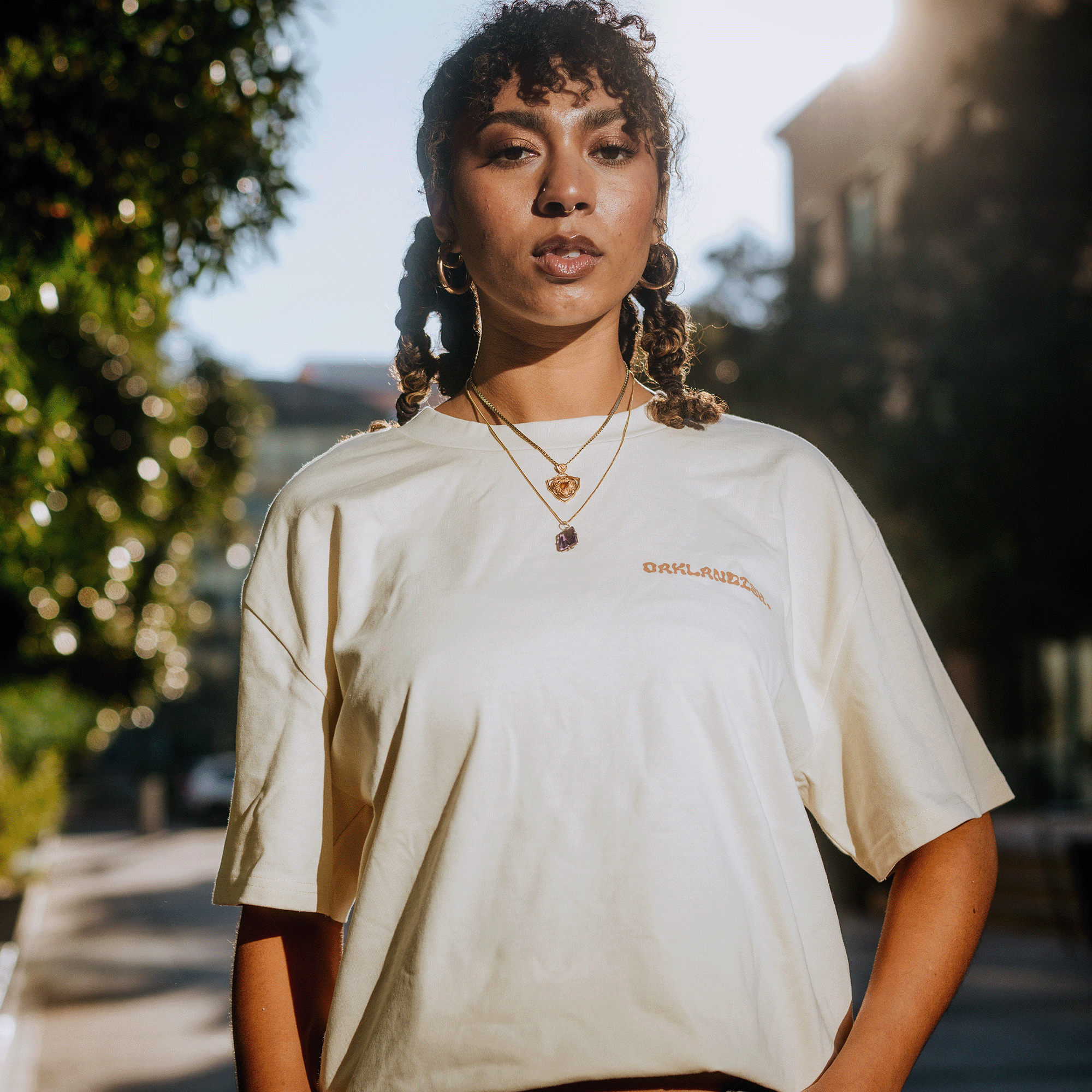 A young woman stands outdoors wearing the Oaklandish "Grow Your Roots" Heavy Tee in a beige boxy fit. Layered necklaces gleam as she gazes confidently at the camera. Her hair is styled in braids, with curly bangs framing her face, while sunlight casts a warm glow over blurred trees and buildings in the background.