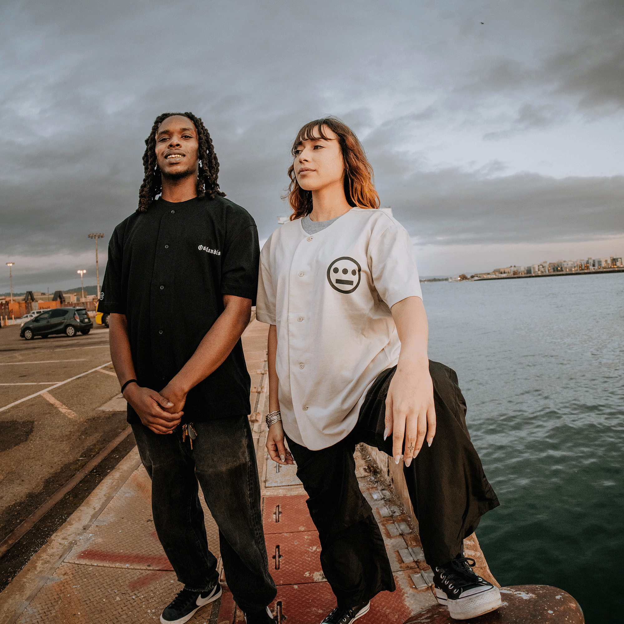 Two individuals pose on a dock beside a body of water, with a cloudy sky and distant shoreline in the background. The person on the left, sporting dreadlocks, is dressed in an Oaklandish Heavy Button-up Baseball Jersey by Oaklandish paired with pants. Meanwhile, the person on the right, with red-brown hair and one foot raised, sports a white shirt featuring a smiley face.