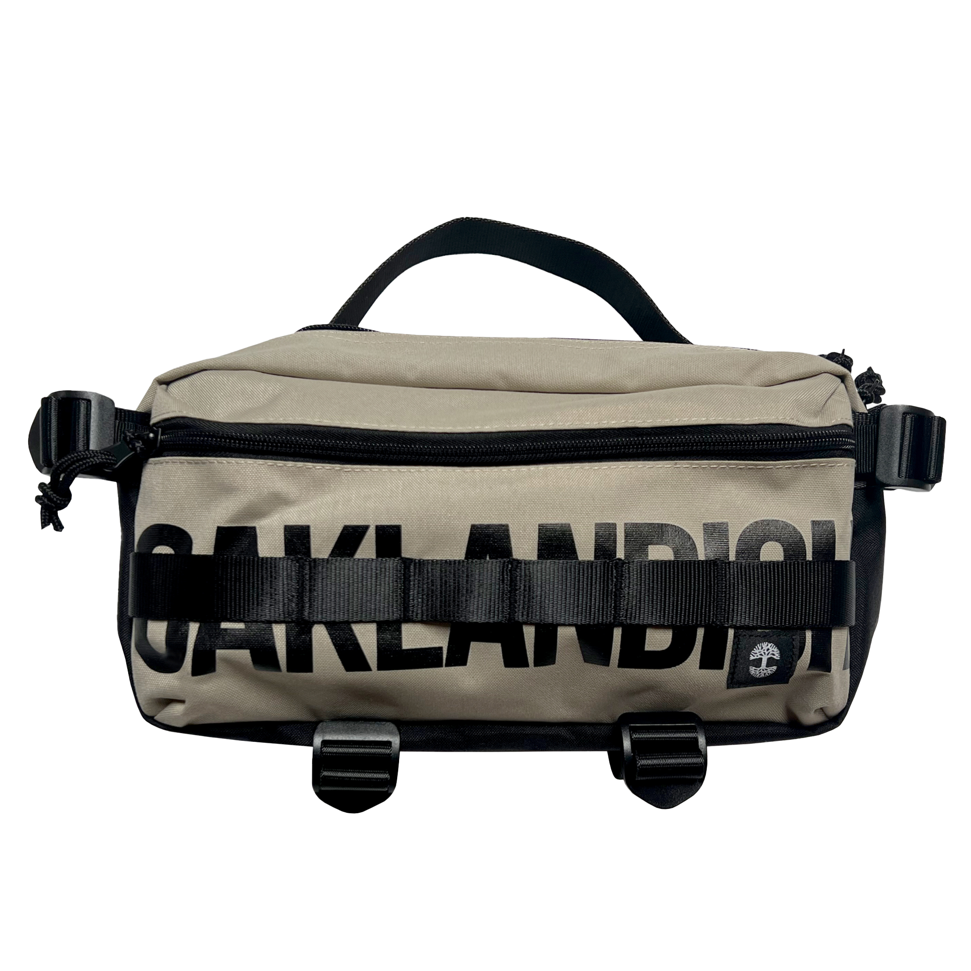 The Oaklandish Hip Bag is a beige and black unisex sling pack with "OAKLANDISH" boldly printed across the front. It features a black handle, buckles, and straps, along with a small circular logo patch on the lower right corner.