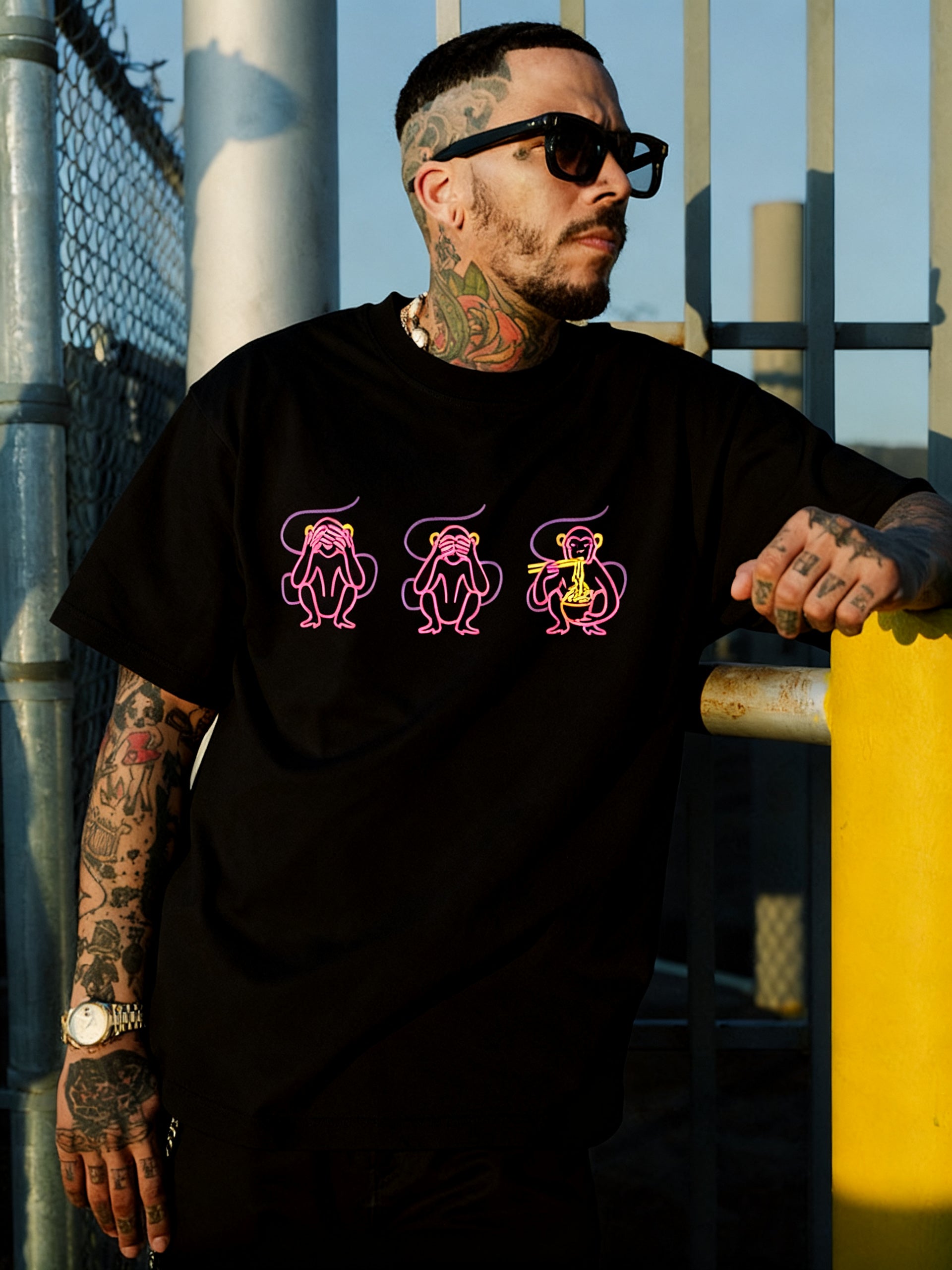A tattooed man in sunglasses and a black Oaklandish Jo's Modern Thai X Oaklandish Tee with neon “see no evil, hear no evil, speak no evil” monkeys stands by a yellow pole and metal fence, looking left in the sunlight.