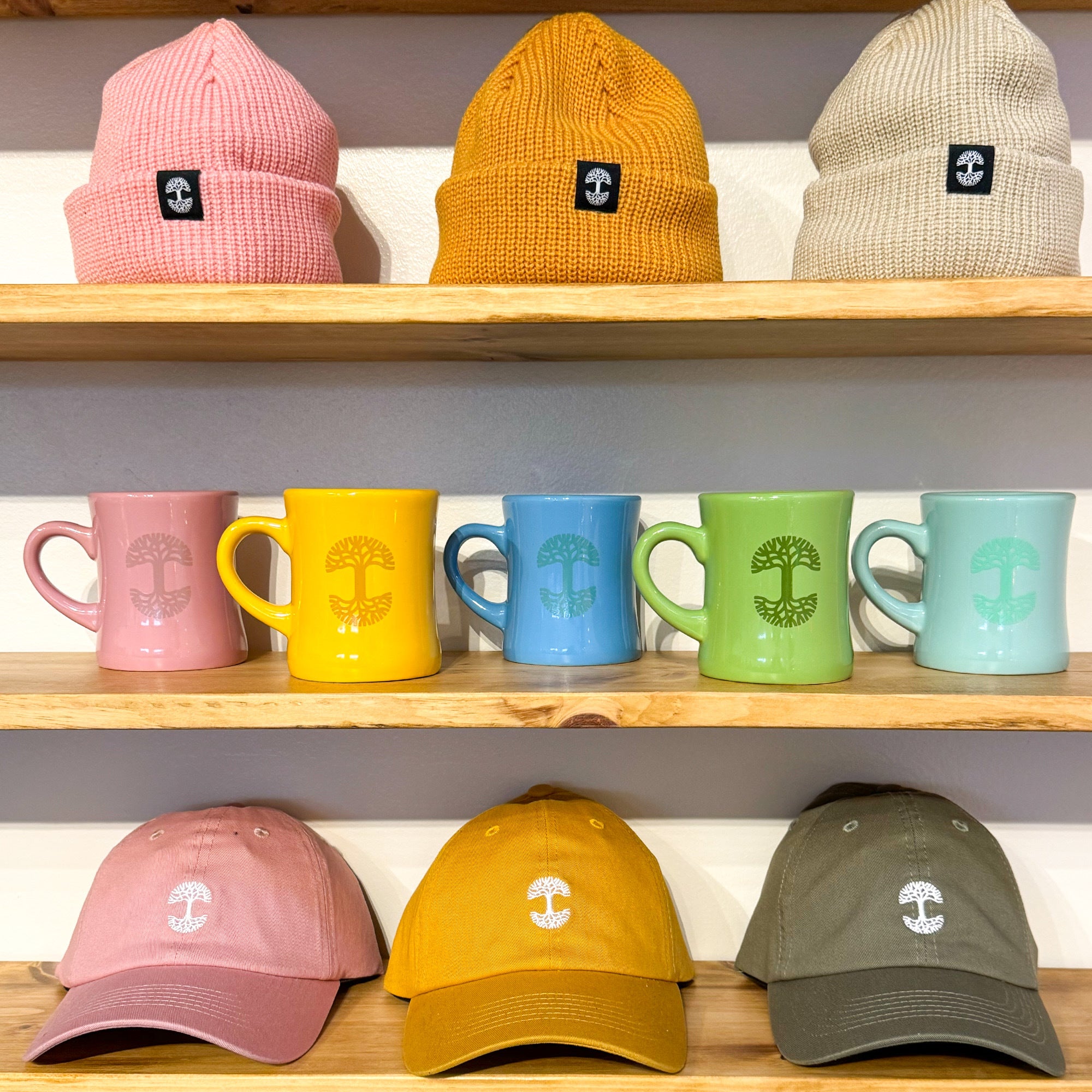 Three wooden shelves showcase various items. The top shelf holds two knitted beanies—one orange and one beige. On the middle shelf, there are four "Oaklandish Diner Mugs" from the brand "Oaklandish," available in pink, yellow, blue, and green. Meanwhile, the bottom shelf displays three baseball caps in pink, yellow, and olive green. Each item prominently features a tree logo.