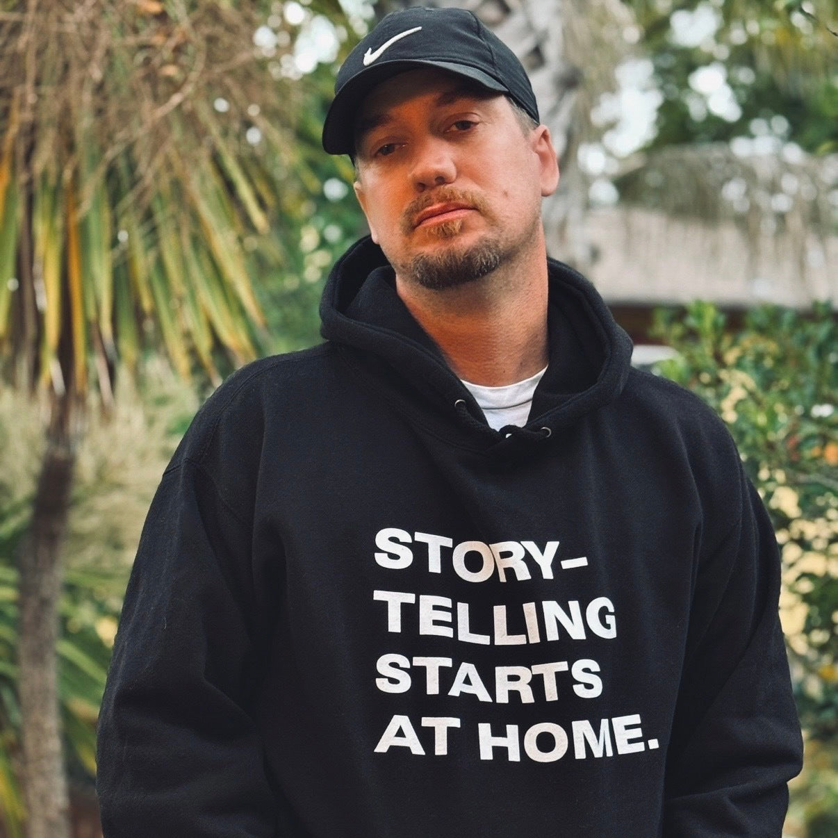 A man in an Oaklandish Make It Bay Hoodie and a black Nike cap stands amid palm trees, representing Bay Area creatives through the Oaklandish collaboration. He faces the camera with a neutral expression.
