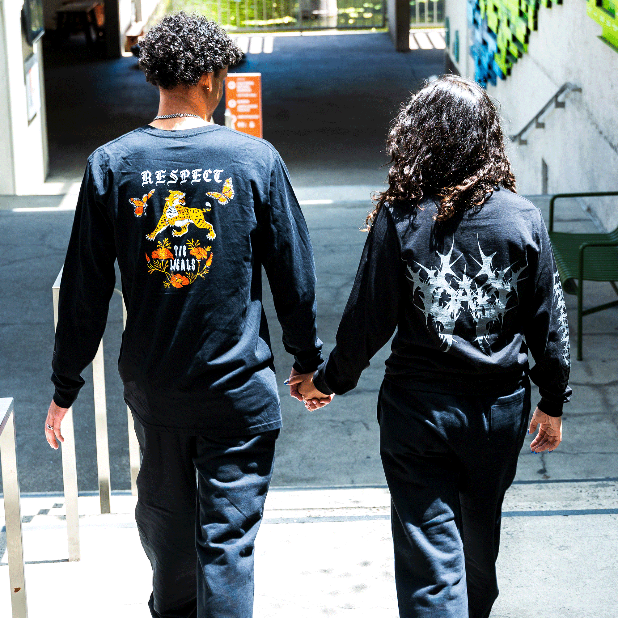 Two people with curly hair, dressed in Oaklandish Cipher LS Tees, walk hand in hand up a sunlit stairway. The person on the left has a shirt featuring colorful designs and "RESPECT" in reflective ink, while the person on the right wears a monochrome version. Green and orange accents decorate the background.