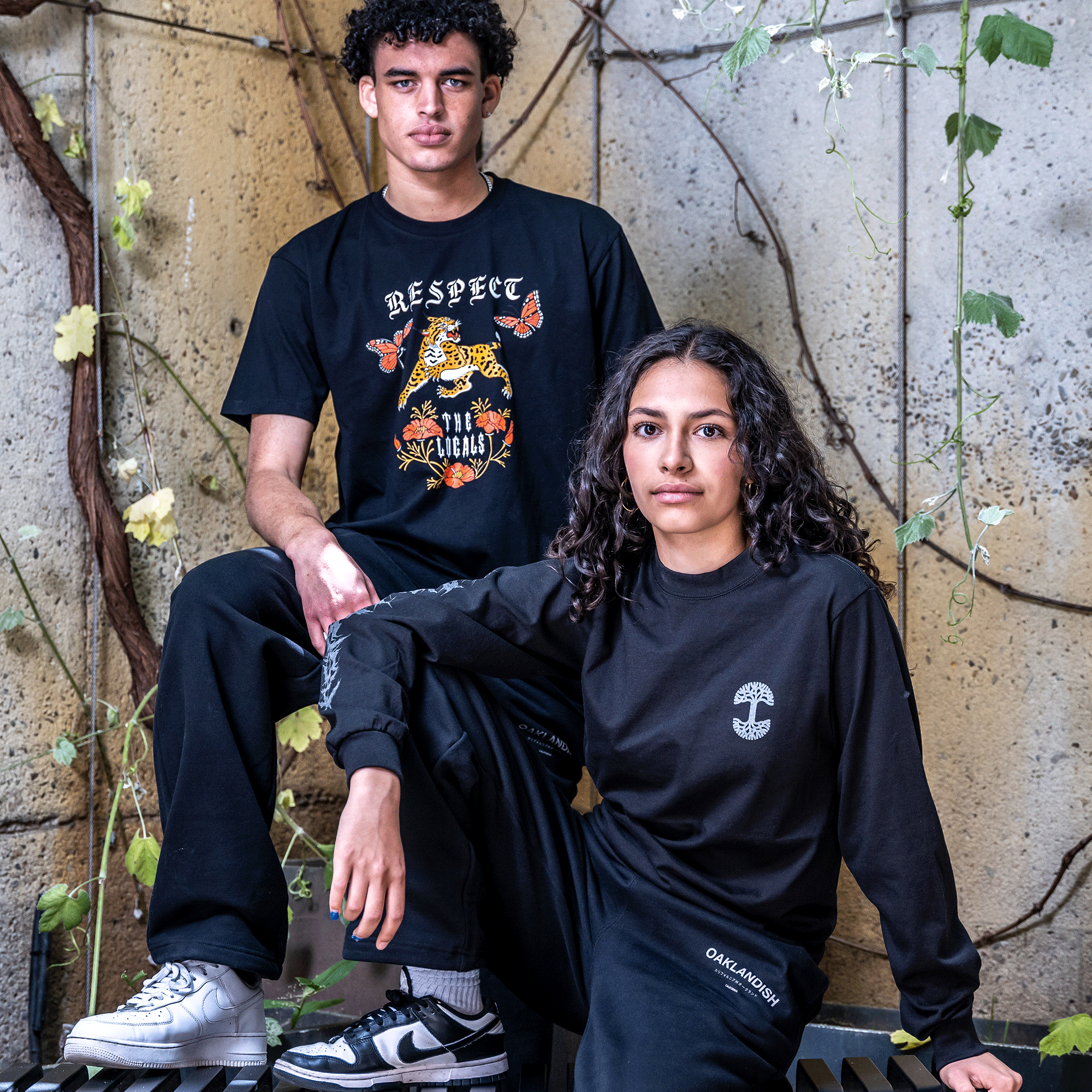 Two people pose confidently in casual streetwear against a rustic urban backdrop with climbing vines. The person on the left wears a black "Respect Originals" T-shirt featuring a vibrant graphic and black pants. The person on the right sports the Cipher LS Tee by Oaklandish, a classic fit long sleeve t-shirt made from 100% cotton with reflective ink lettering.