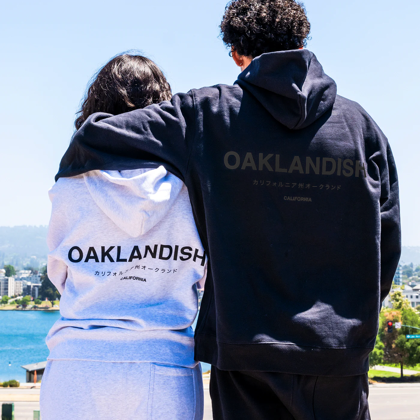 A person wearing a light gray Modern Type hoodie by Oaklandish is hugging another individual in a black unisex version of the same hoodie. Both hoodies feature the word "OAKLANDISH" on the back. They stand facing away from the camera, in front of a body of water with a clear sky and trees in the background.