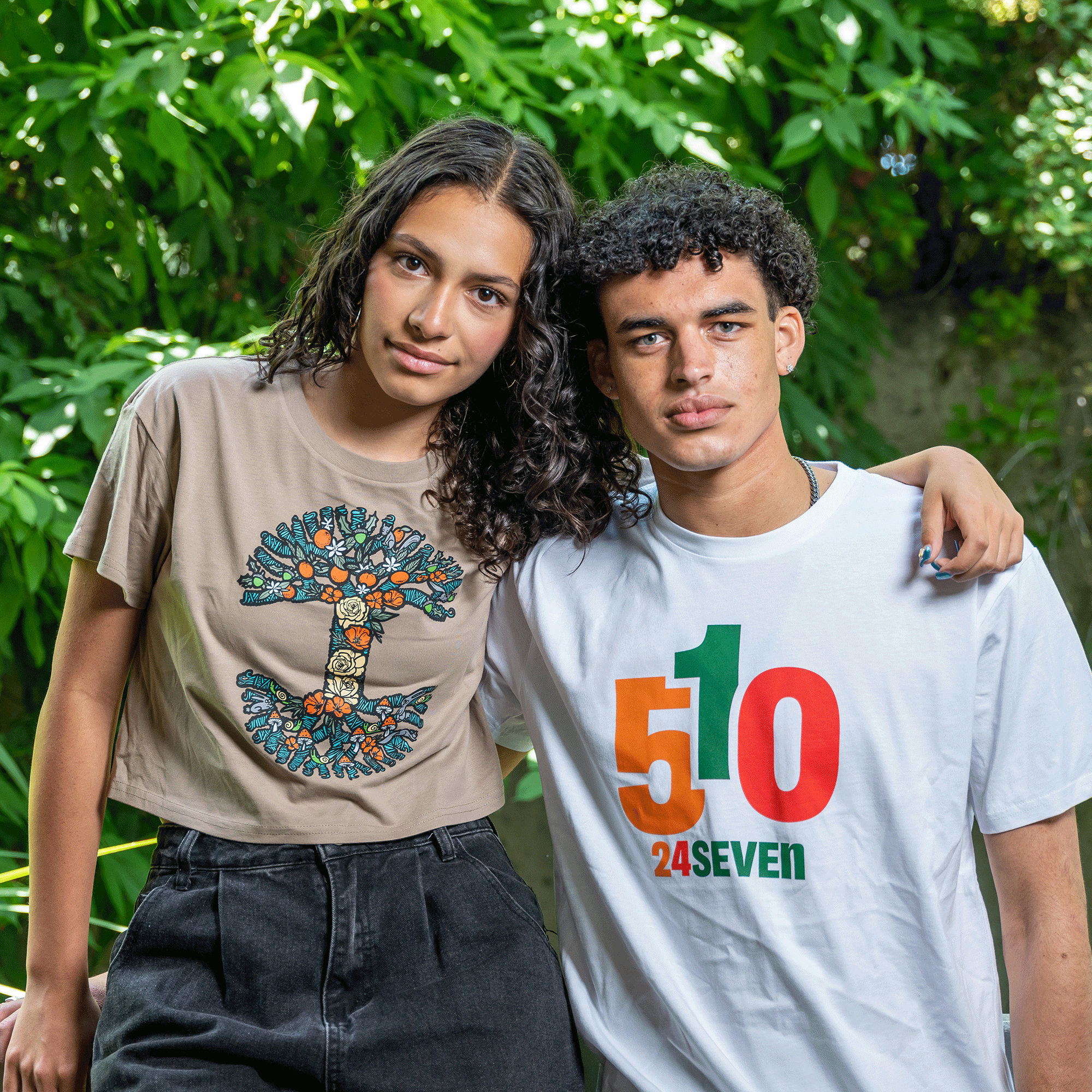 Two young individuals stand close together outdoors, posing for the camera. The person on the left wears an Oaklandish Women's Thriving Tree Crop Tee in beige featuring a classic tree logo, while the person on the right dons a white T-shirt with "510" in orange, green, and red alongside the text "24SEVEN." Lush greenery is in the background.