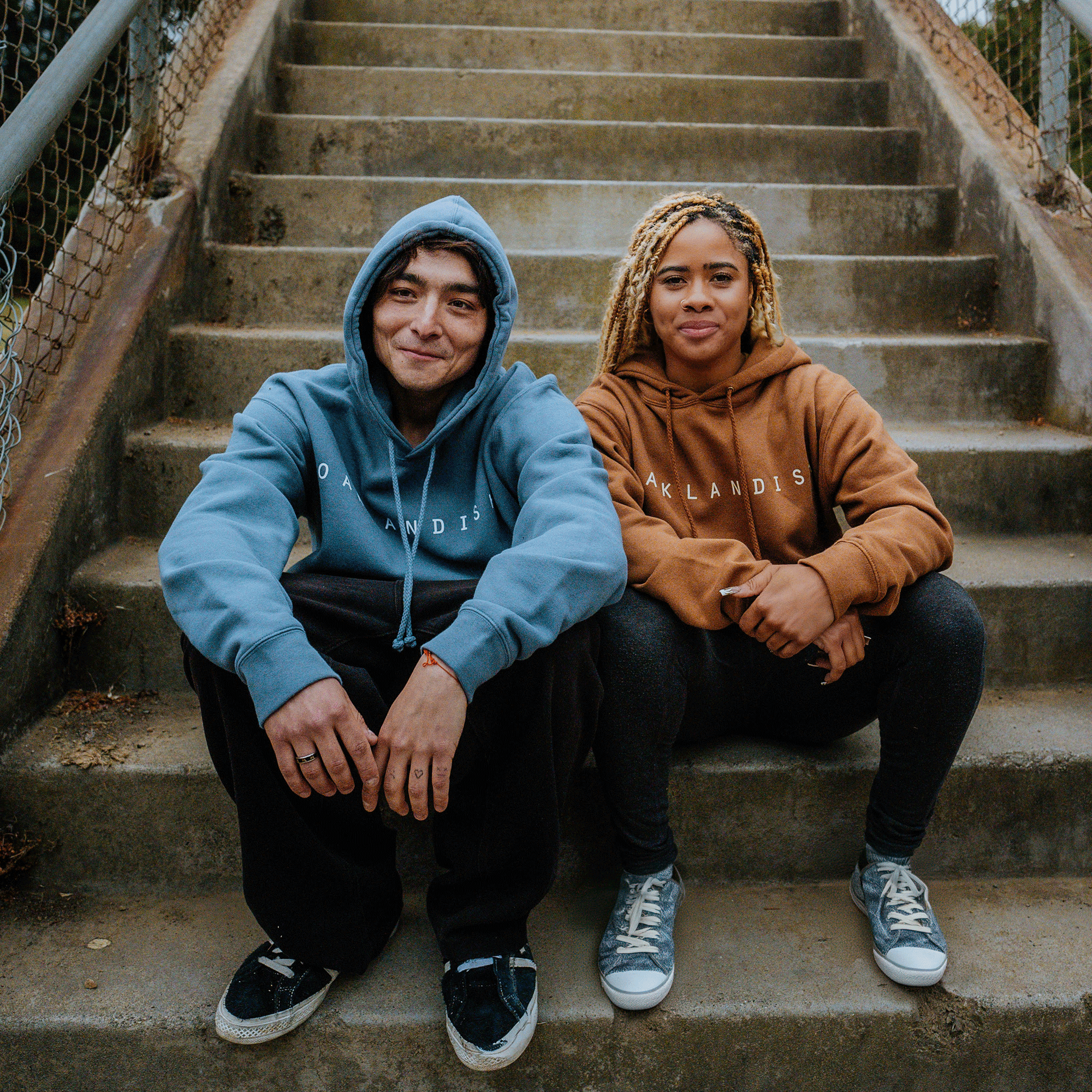 Two people sit side by side on outdoor concrete steps, both showcasing their Oaklandish pride in pullover hoodies adorned with the "OAKLANDISH" logo. The person on the left sports a storm blue Oaklandish Classic Hoodie and black sneakers, while the one on the right wears a brown Oaklandish Classic Hoodie paired with gray sneakers. They exude a relaxed and friendly demeanor.