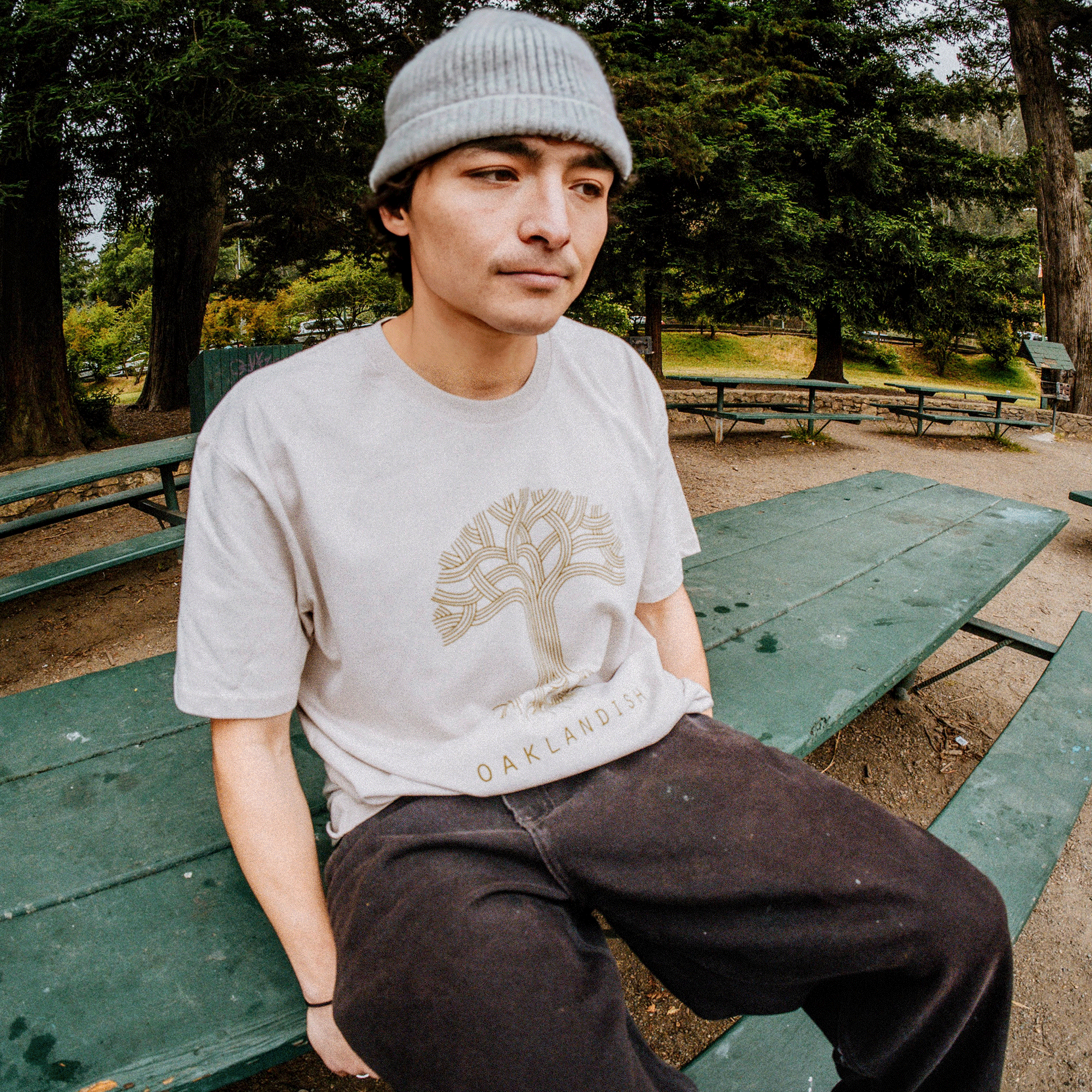 A person wearing a gray beanie and an "Oaklandish Classic Logo Tee" from Oaklandish sits on a green picnic table in a park. The background features tall trees and green foliage. The person has a thoughtful expression and casual style with dark pants.