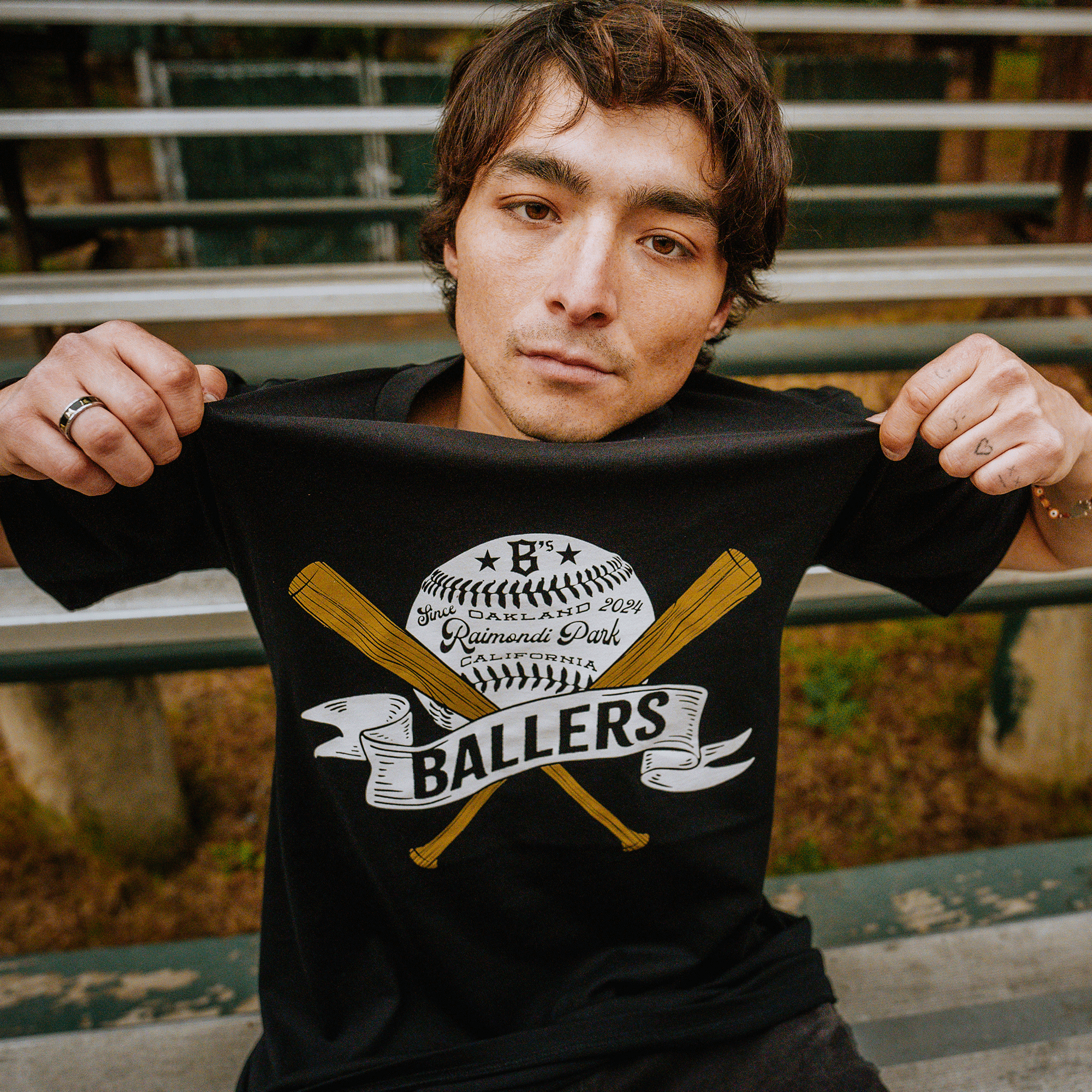 A person with short brown hair is seated on bleachers, holding up the Oakland Ballers "A New Era of Oakland Baseball Tee" with both hands. The black T-shirt features a design of a baseball, two crossed bats, a ribbon, and the words "B's Portland 2019, Rainwater Park, Oakland Ballers." The person is looking directly at the camera.