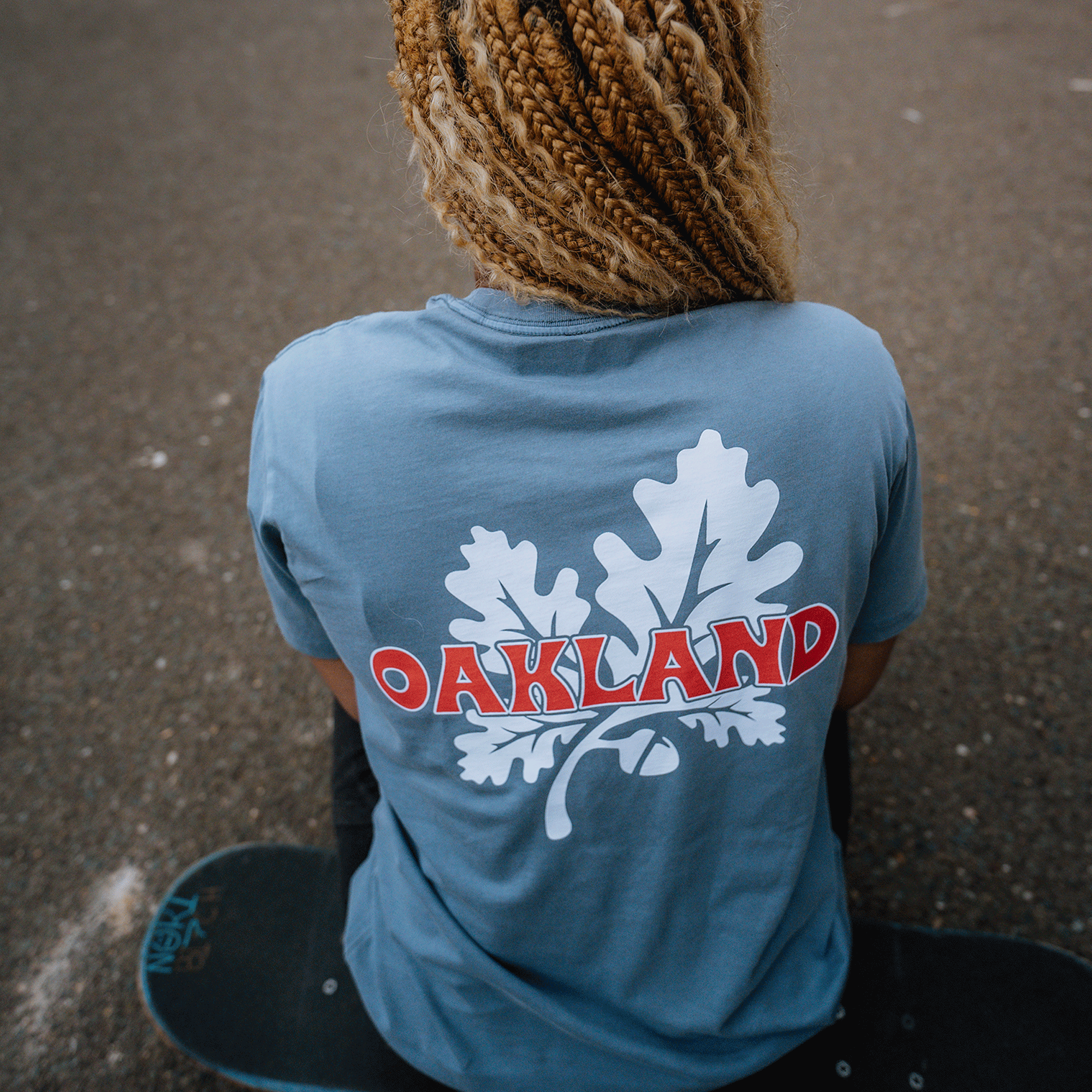 A person with blonde braided hair sits on a skateboard facing away from the camera, sporting the Oaklandish Grocery Run Tee. This fresh blue classic fit t-shirt, made of 100% cotton, features a large white oak leaf graphic and the word "OAKLAND" in bold red letters outlined in white. The background is an asphalt surface.