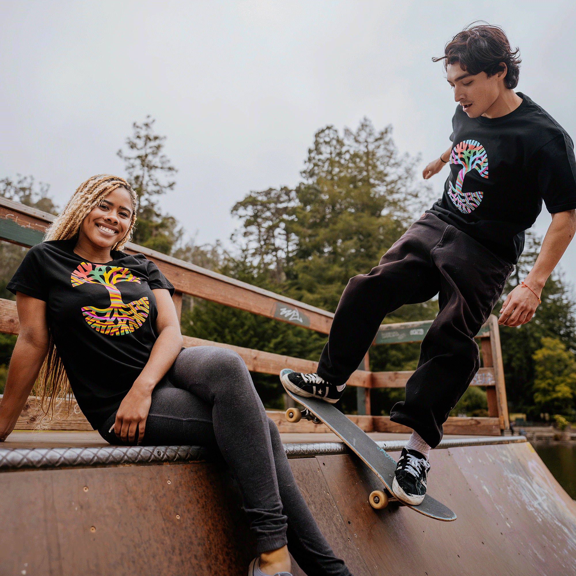 A young man and woman are at a skate park. The woman, with long braided hair, sits on the edge of a ramp smiling, while the man skateboards beside her. Both are wearing Oaklandish Stripe Tree Tees featuring colorful abstract designs and made from 100% cotton. Trees and a cloudy sky are visible in the background.