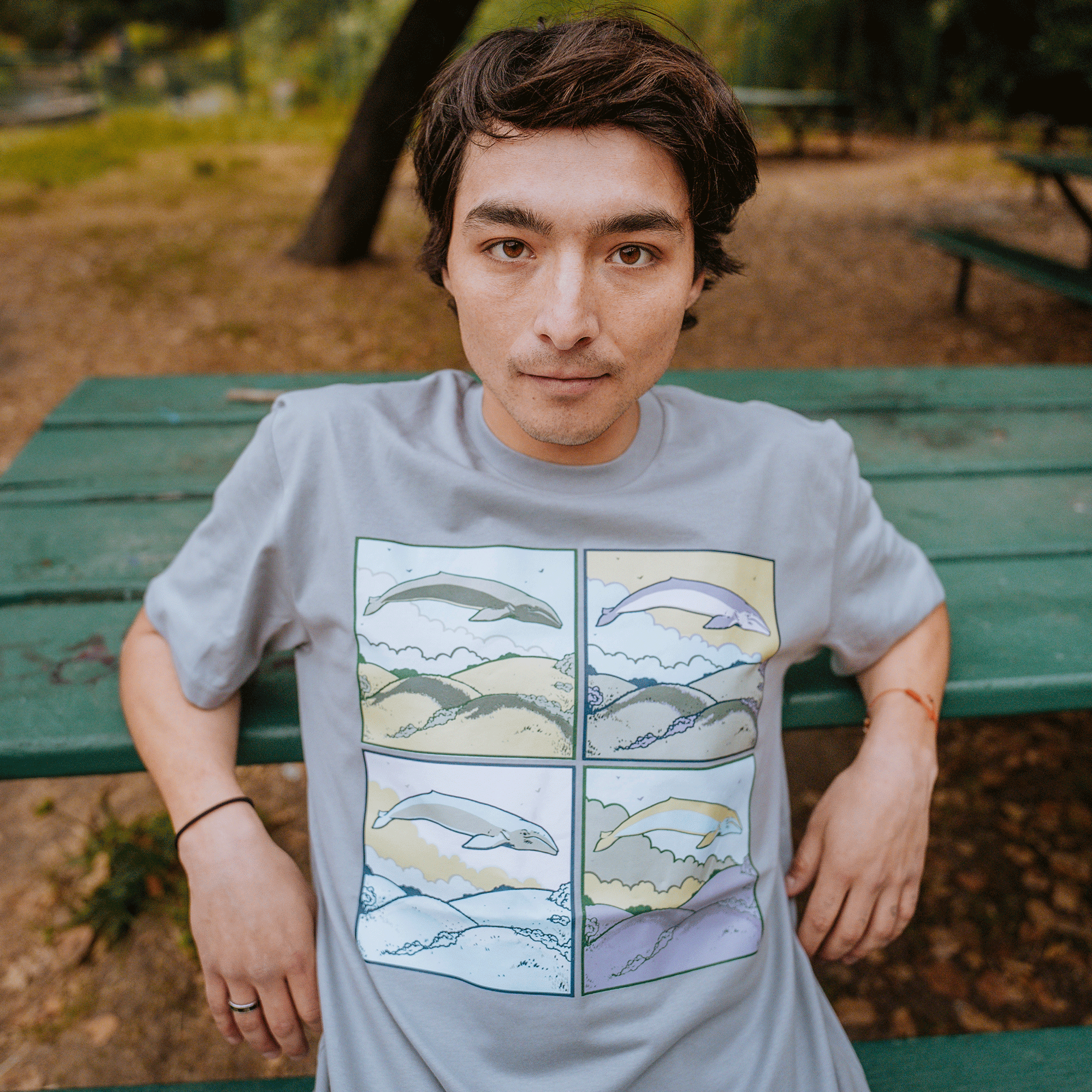 A person with medium-length hair, wearing the Whale Dreams Tee from Oaklandish men's collection—featuring a light gray, 100% cotton fabric and a four-panel design of a whale in various natural settings—sits on a green wooden picnic table in an outdoor park. The scene is framed by dirt ground, trees, and another picnic table as the person looks directly at the camera.