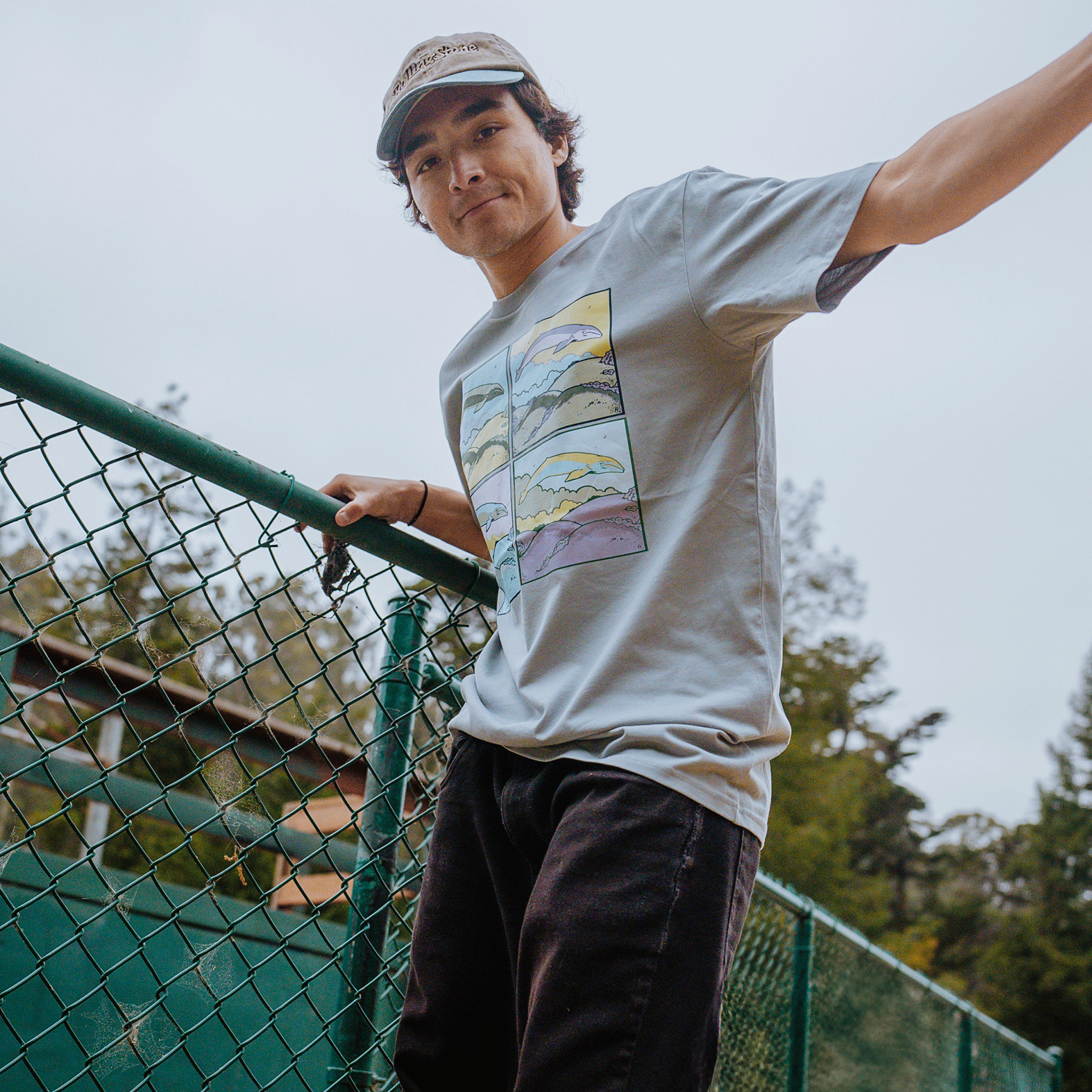 A young man with dark hair wearing a cap and a light gray, 100% cotton, classic fit Whale Dreams Tee by Oaklandish, featuring four colorful mountain illustrations, stands next to a green chain-link fence. He has one arm resting on the fence while the backdrop showcases trees and an overcast sky—part of our latest men's collection.