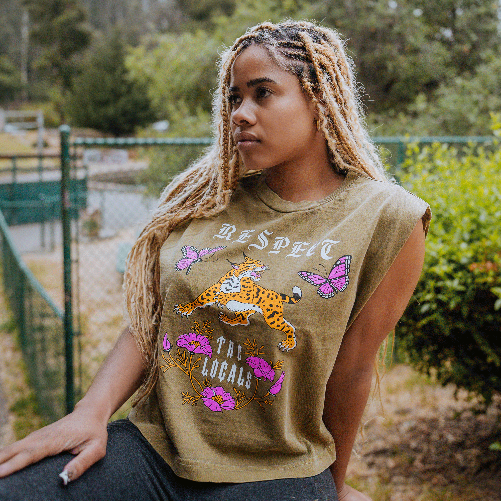 A person with long, braided blonde hair is outdoors, leaning on a ledge. They are wearing the Women's Respect the Locals Tank by Oaklandish, a sleeveless olive-green top made of 100% cotton featuring tattoo-style graphic prints of a cheetah, purple flowers, and butterflies along with the words "RESPECT THE LOCALS." The background showcases a chain-link fence and greenery.