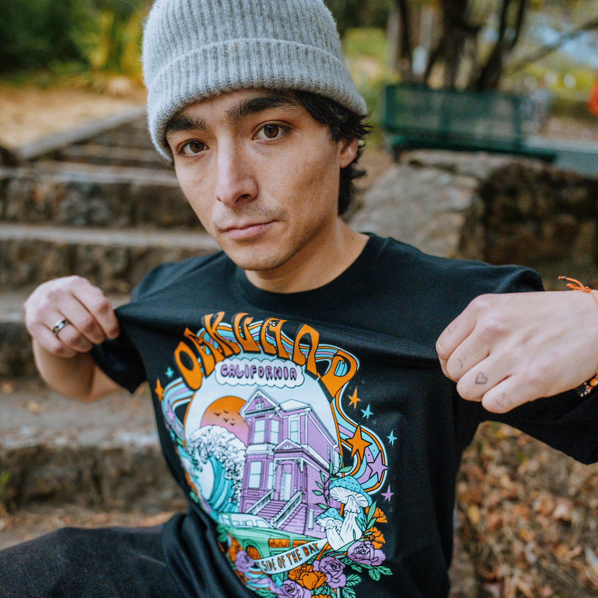 A person stands outdoors, wearing a gray beanie and holding up the front of the Oakland Dream Tee from Oaklandish. The classic fit black T-shirt, crafted from 100% cotton, showcases a colorful design featuring the word "OAKLAND" prominently displayed alongside images of a house, waves, and vibrant graphics. Stone steps and greenery are visible in the background.