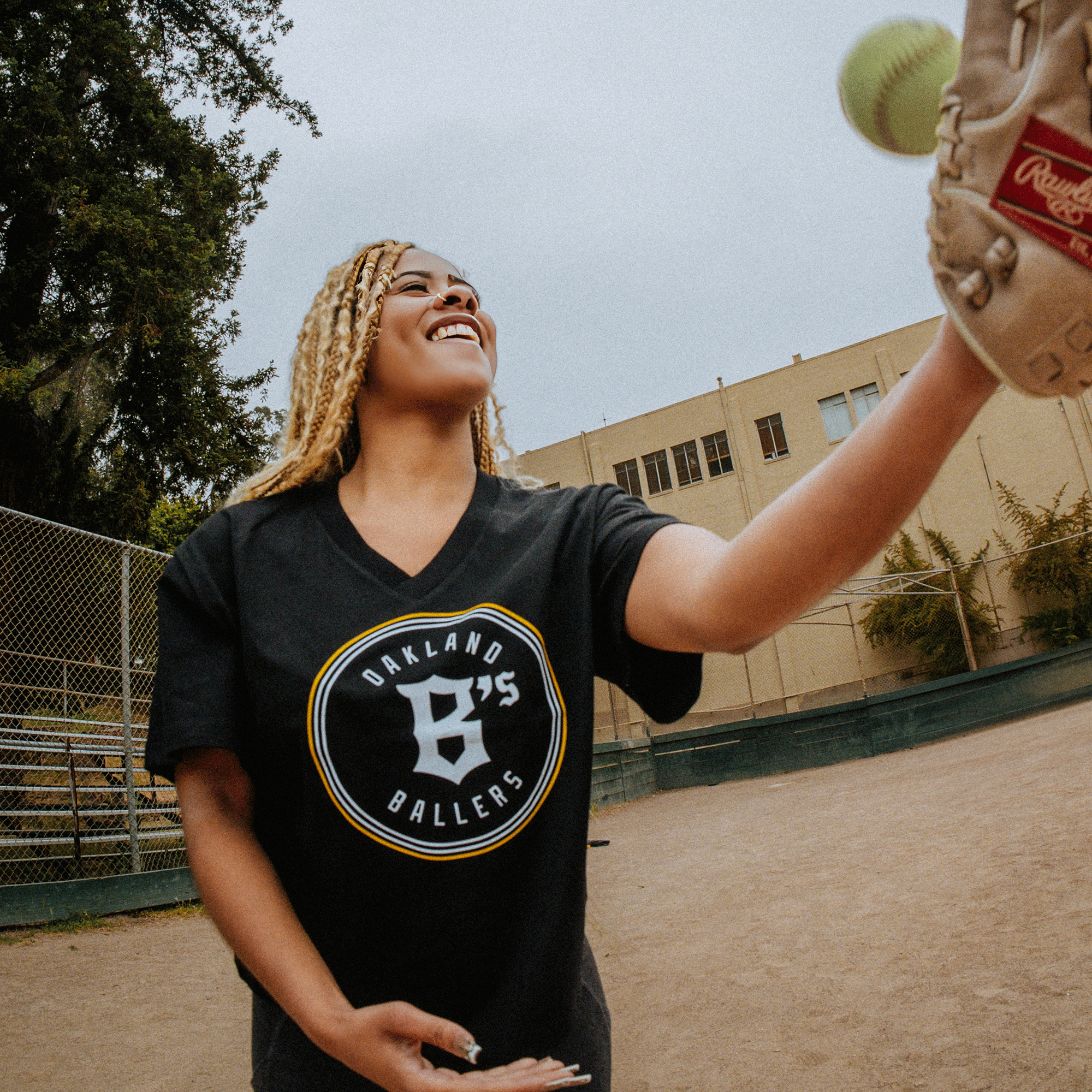 A smiling woman with braided blonde hair tosses a softball in the air while wearing a black "Women's Oakland Ballers Logo Relaxed V Neck Tee" from Oakland Ballers. She stands in a dirt baseball field with a school building and chain-link fence in the background, representing East Bay pride. She has a softball glove on her left hand.