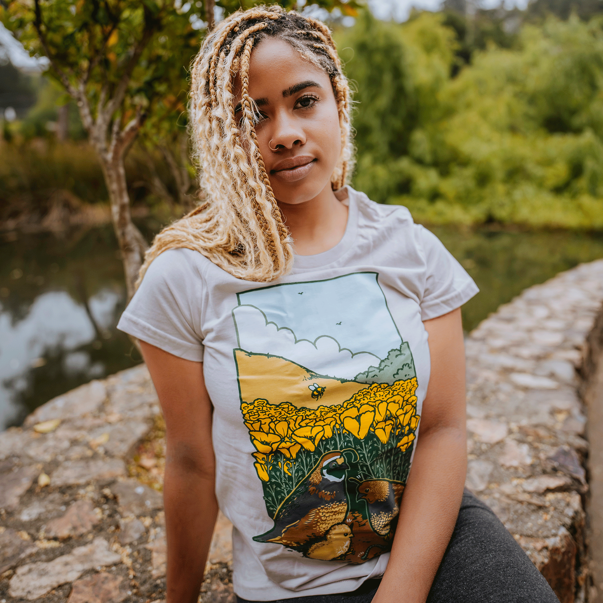 A young woman with long, blonde, curly hair sits on a stone ledge by a tranquil pond surrounded by greenery. She wears the Oaklandish Women's Quail Family Tee, a classic fit, 100% cotton women's t-shirt featuring an illustration of a vibrant field of yellow flowers with ducks at the forefront and a landscape in the background.