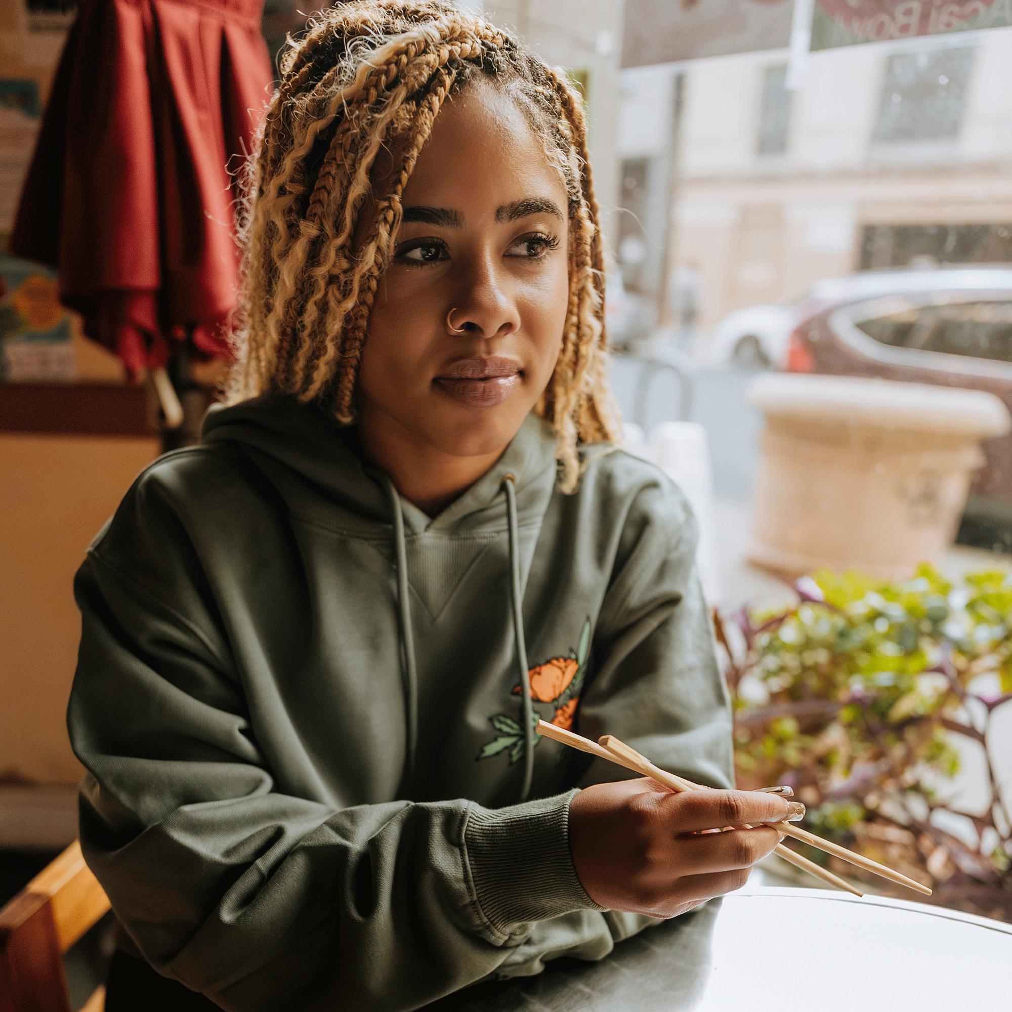 A person with blonde dreadlocks sits at a table in a cozy cafe, holding a pair of chopsticks. They wear the Oakland Dream Premium Hoodie by Oaklandish, featuring a small flower design made from luxe French terry. Sunlight streams through the window, illuminating their thoughtful expression. The background includes a street view with a parked car and plants.