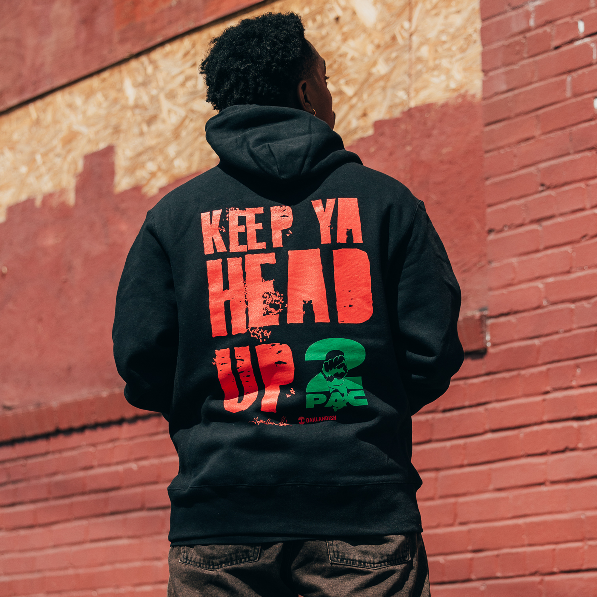 A person stands with their back to the camera wearing the "You Got To KYHU Hoodie" by Oaklandish. This unique Oaklandish x Tupac collaboration features bold red text that reads "KEEP YA HEAD UP," complemented by green and red graphics against a brick wall background, encapsulating a motivational message deeply rooted in Oakland culture.