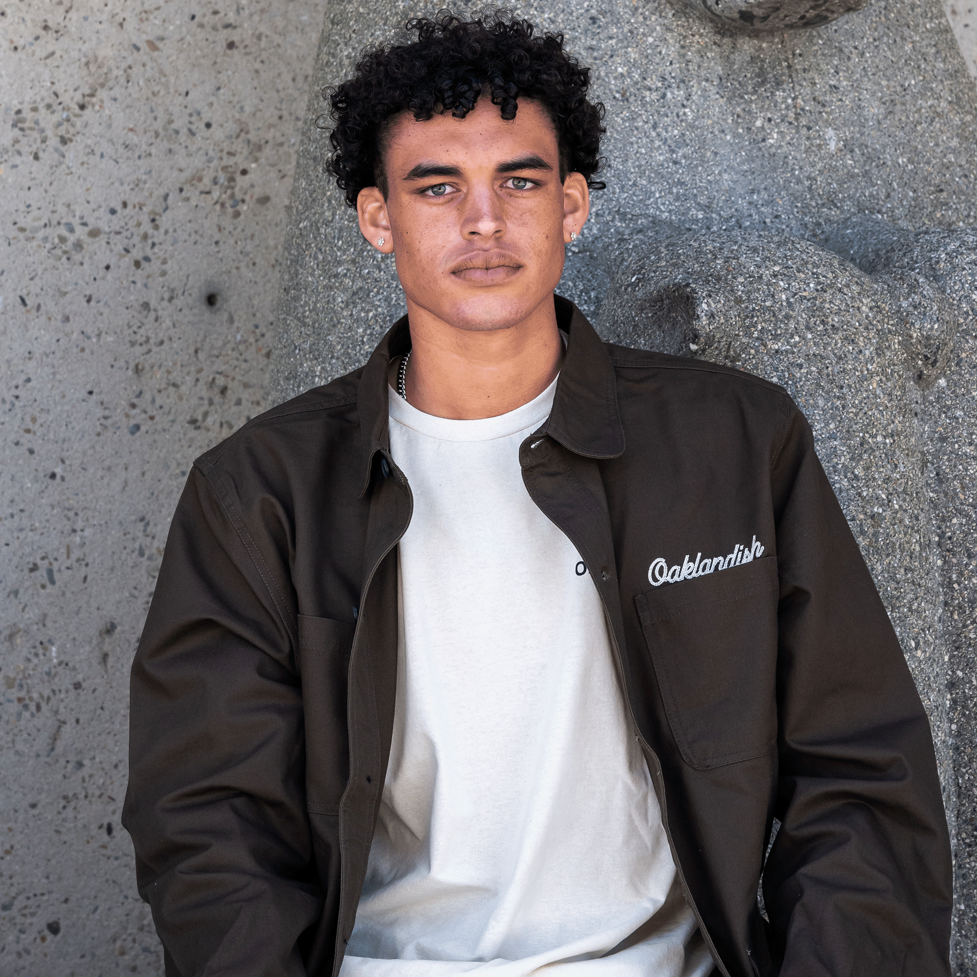 A person with curly dark hair and a stubble beard is seated against a textured concrete wall. They are wearing the Forge Field Jacket, a dark brown collared shirt jacket from Oaklandish, over a white t-shirt with "Oaklandish" written on the left side. Their expression is neutral, and they are looking directly at the camera.