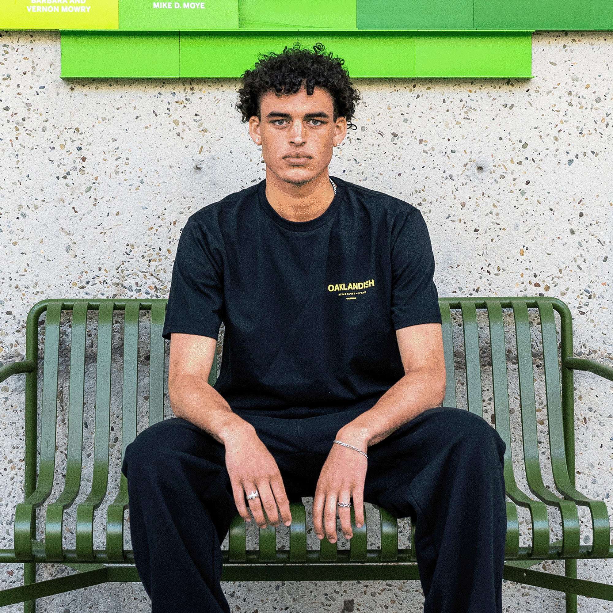 A young man with curly hair is sitting on a green metal bench, staring directly at the camera. He is wearing a black Modern Type Tee by Oaklandish, made from 100% cotton and featuring the word "Oaklandish" printed in yellow on the chest. He pairs it with black pants and several rings. The background features a textured wall with green and yellow name plates.