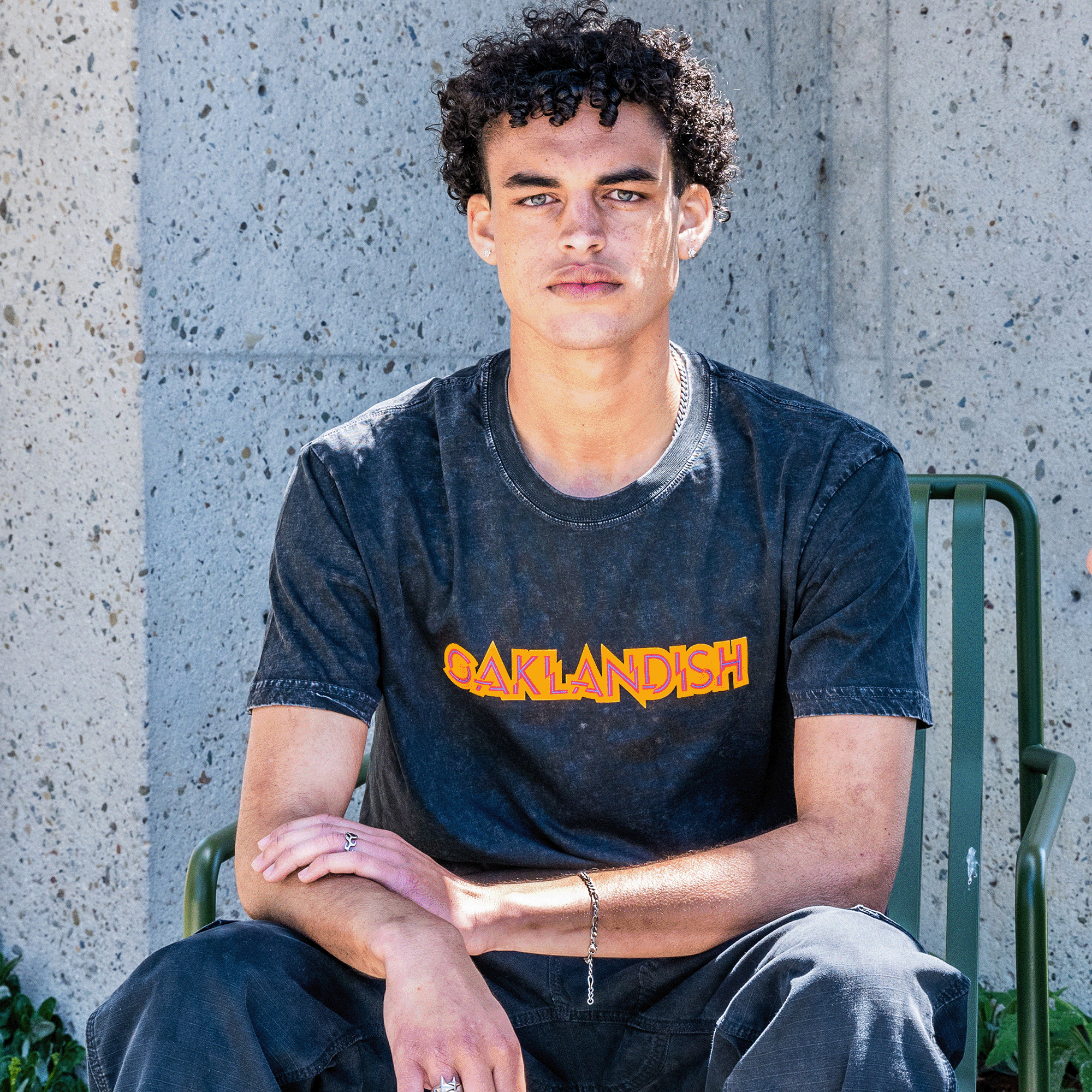 Male model wearing stone cotton t-shirt with Oaklandish wordmark in orange and red.