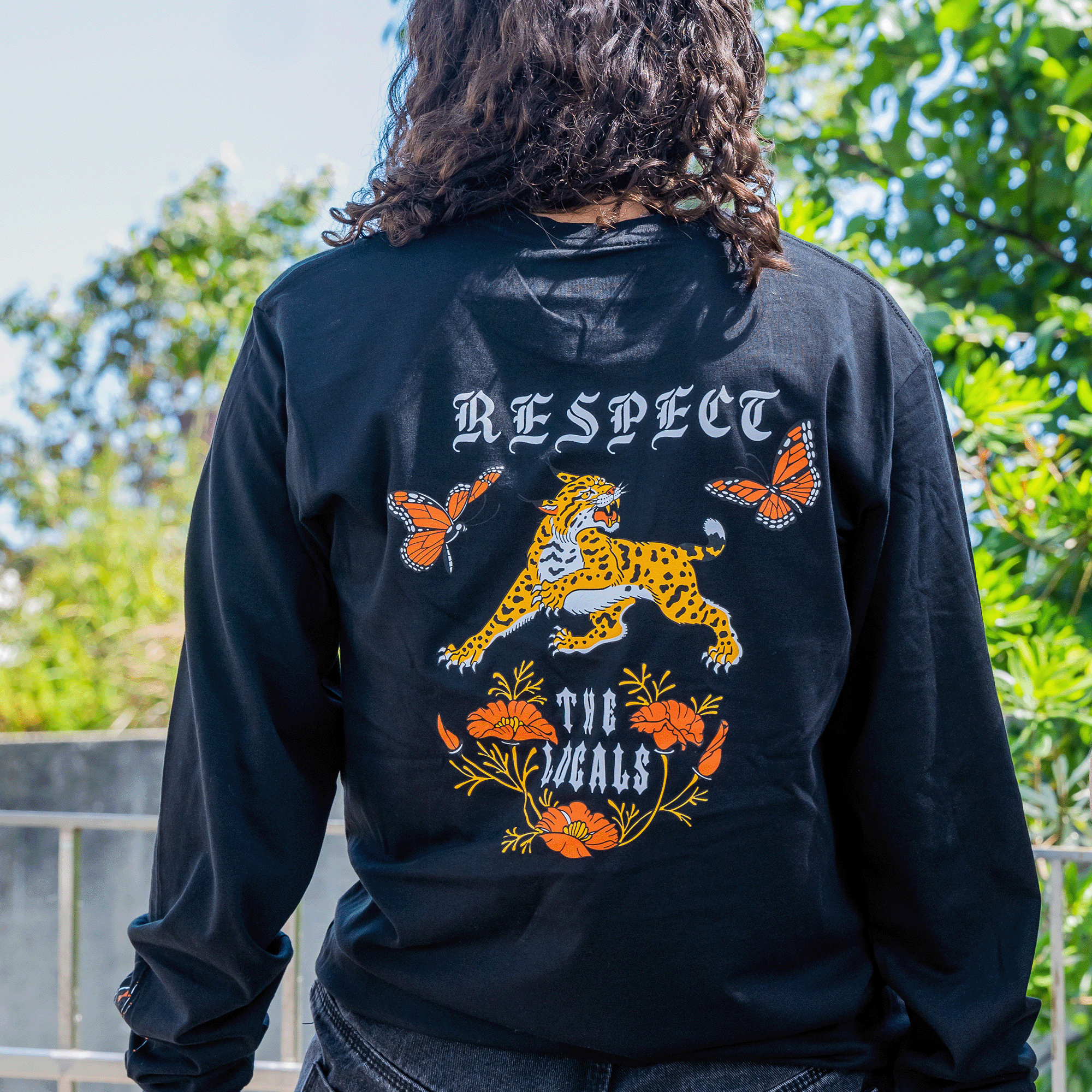 A person stands outdoors with their back to the camera, wearing the Oaklandish Respect the Locals LS Tee. The black long-sleeve t-shirt, made of 100% cotton, showcases a colorful design featuring a leopard, two orange butterflies, orange flowers, and the words "RESPECT THE LOCALS." Lush greenery is blurred in the background.