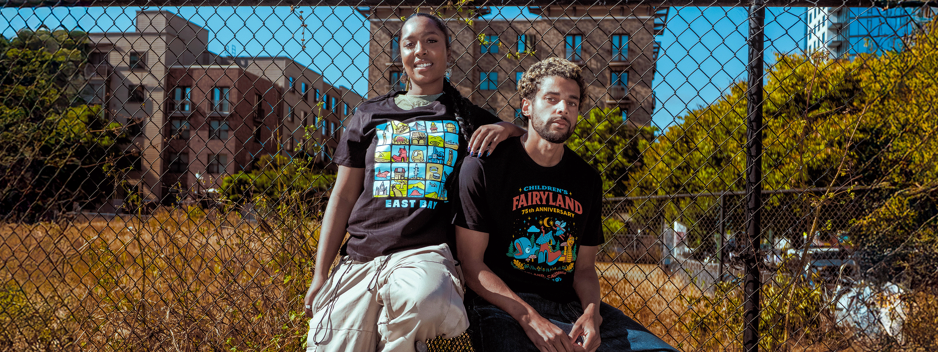 Two people sitting behind a chain-link fence with a cityscape in the background
