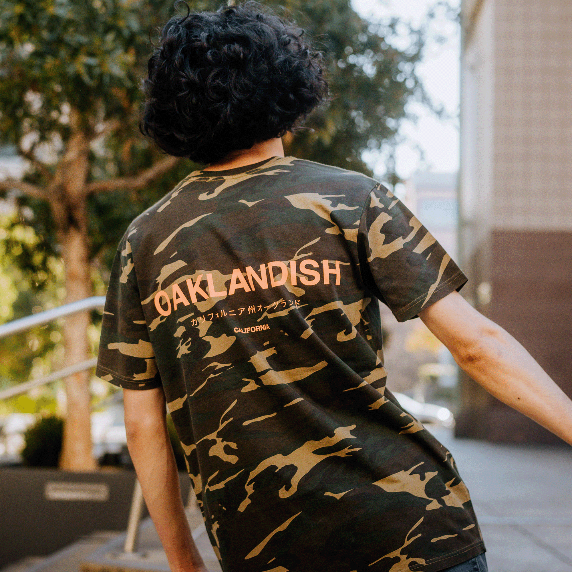 A person with curly hair is wearing a classic fit, 100% cotton Modern Type Tee from Oaklandish. This camouflage t-shirt features "Oaklandish" in large letters on the back, along with stylized Japanese characters and "California" printed below. They are outdoors, surrounded by trees and a building in the background.