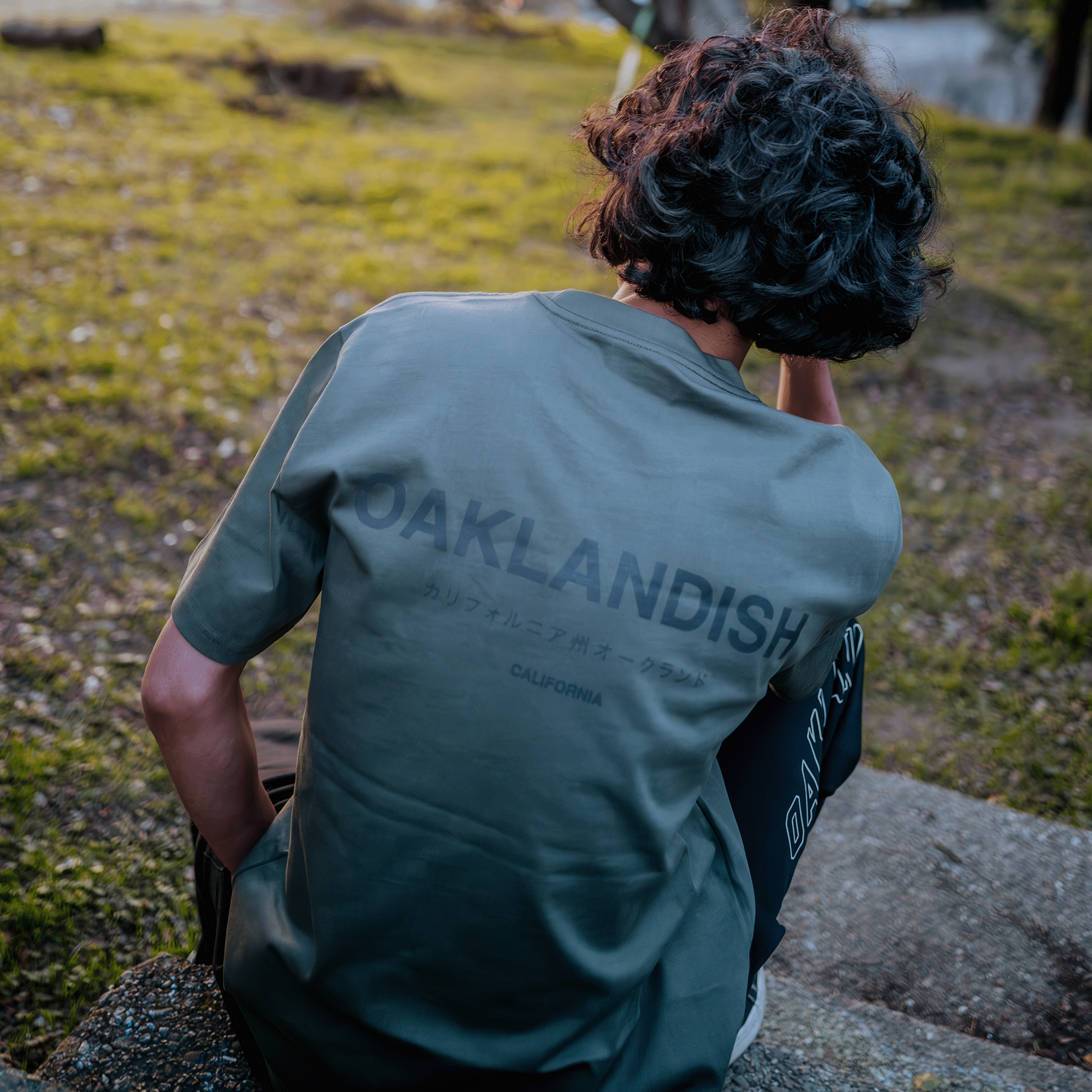 A person with wavy hair sits outdoors on stone steps, facing away. They wear a heavyweight cotton dark green Modern Type Heavy Tee from Oaklandish, featuring "OAKLANDISH CALIFORNIA" printed on the back. The background includes grassy areas and a blurred tree, hinting at a park or natural setting in soft lighting.