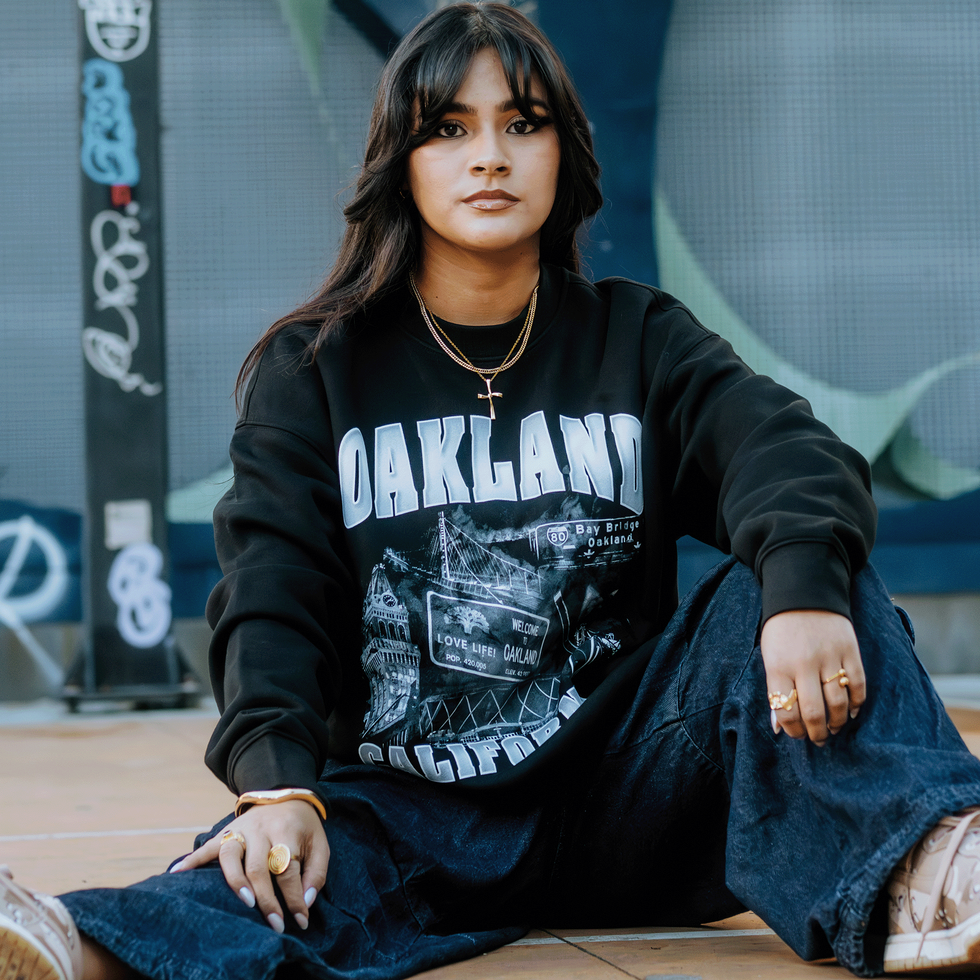 A person with long dark hair sits on the ground wearing a black "Montage Crew" sweatshirt by Oaklandish and wide-leg jeans. Adorned with layered gold necklaces and rings, they complete the look with white sneakers. The graffiti-covered wall and wooden floor backdrop highlight their calm, serious expression.