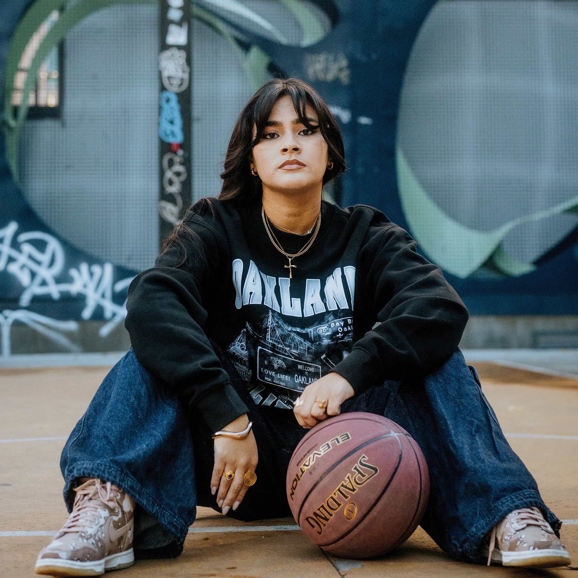 A person wearing the Montage Crew sweatshirt by Oaklandish and jeans sits on a basketball court with a basketball in front. They have long dark hair adorned with hoop earrings and a cross necklace. The vibrant background features graffiti and a striking blue abstract mural.