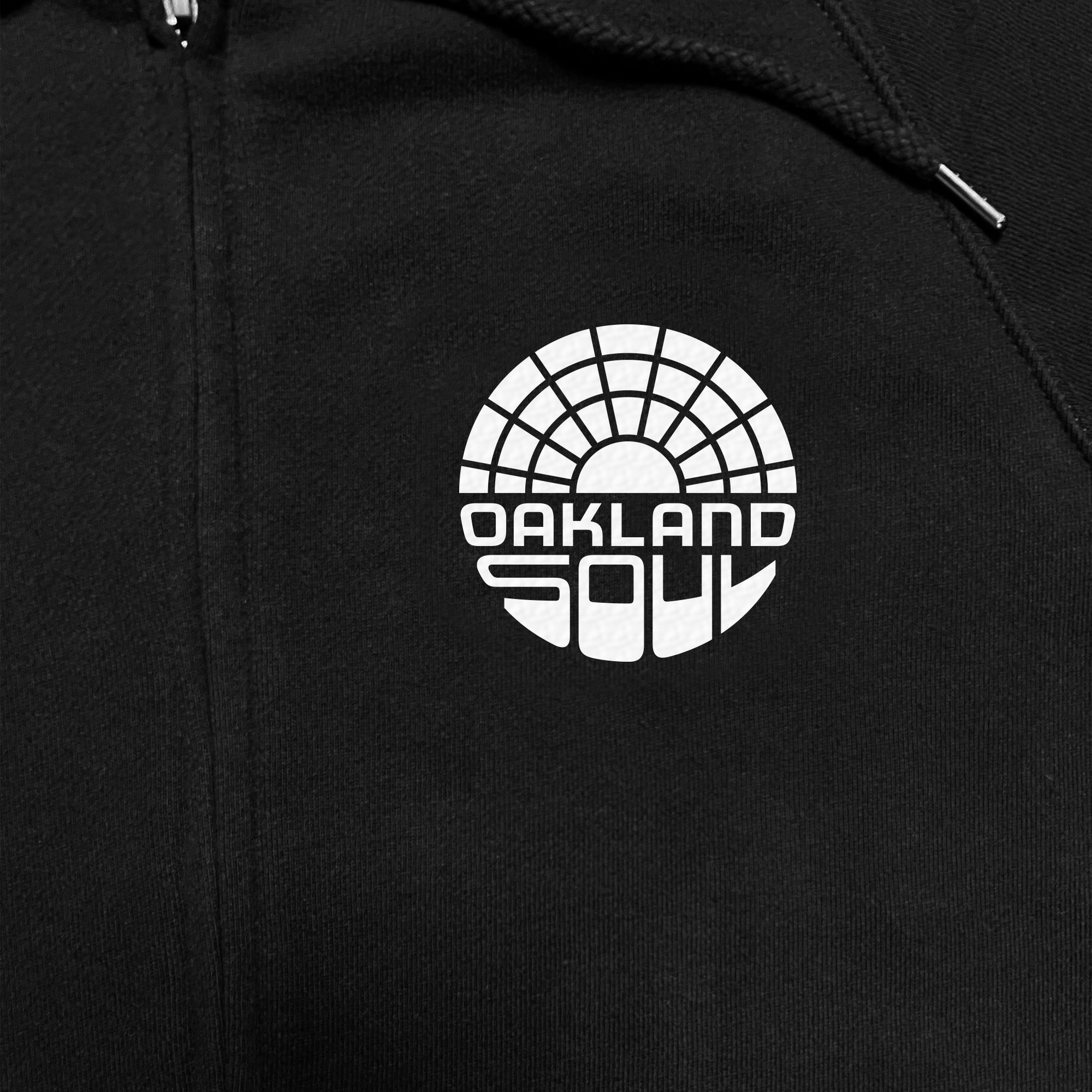 Close-up image of the Oakland Soul Logo Zip, a black unisex zip hoodie from Oakland Roots SC. The hoodie features a white "Oakland Soul" logo on it, symbolizing social good with a semi-circular, mosaic-like design above the text. The words "OAKLAND" and "SOUL" are written in bold, blocky letters, with "OAKLAND" appearing above "SOUL".