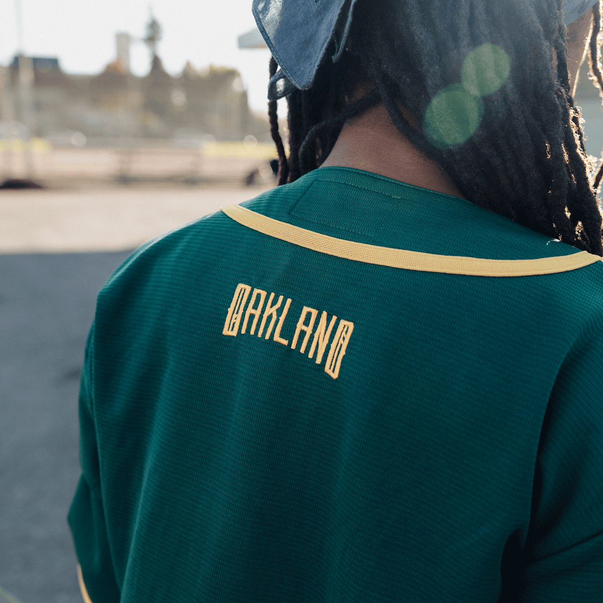 A person with dark locs seen from the back wears a forest green Oakland Button Up Jersey with yellow trim, featuring “OAKLAND” embroidered in yellow block lettering across the upper back against a softly blurred outdoor setting.