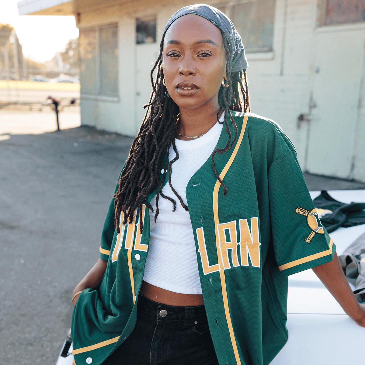 A woman with long dreadlocks and a bandana leans against a white car wearing an unbuttoned forest green Oakland Button Up Jersey over a white cropped tank top, featuring yellow lettering and a gold baseball bat emblem on the sleeve.
