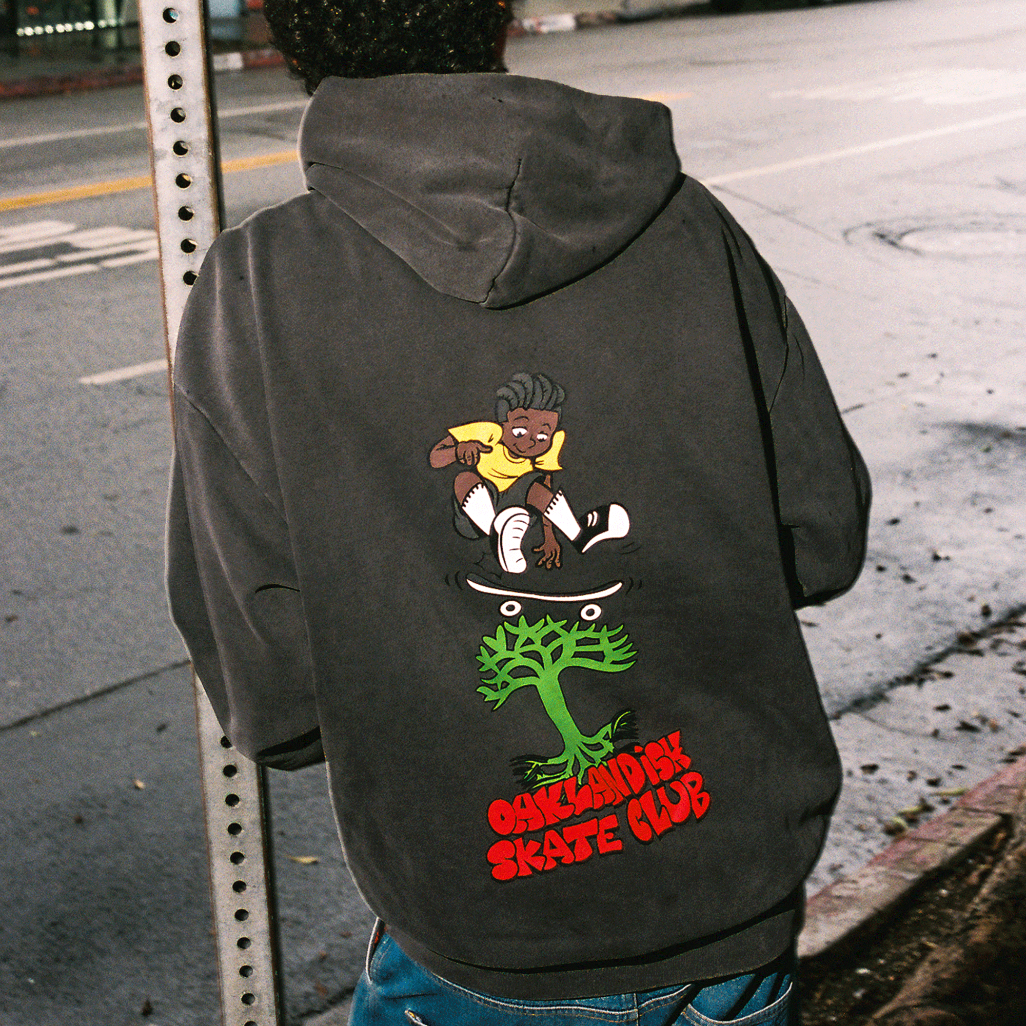 A person with curly hair wears the Oaklandish Skate Club Hoodie, featuring a cartoon skater tricking over a green tree and red “Oaklandish Skate Club” text on the back. They stand on an empty street under cloudy skies. Brand: Oaklandish.