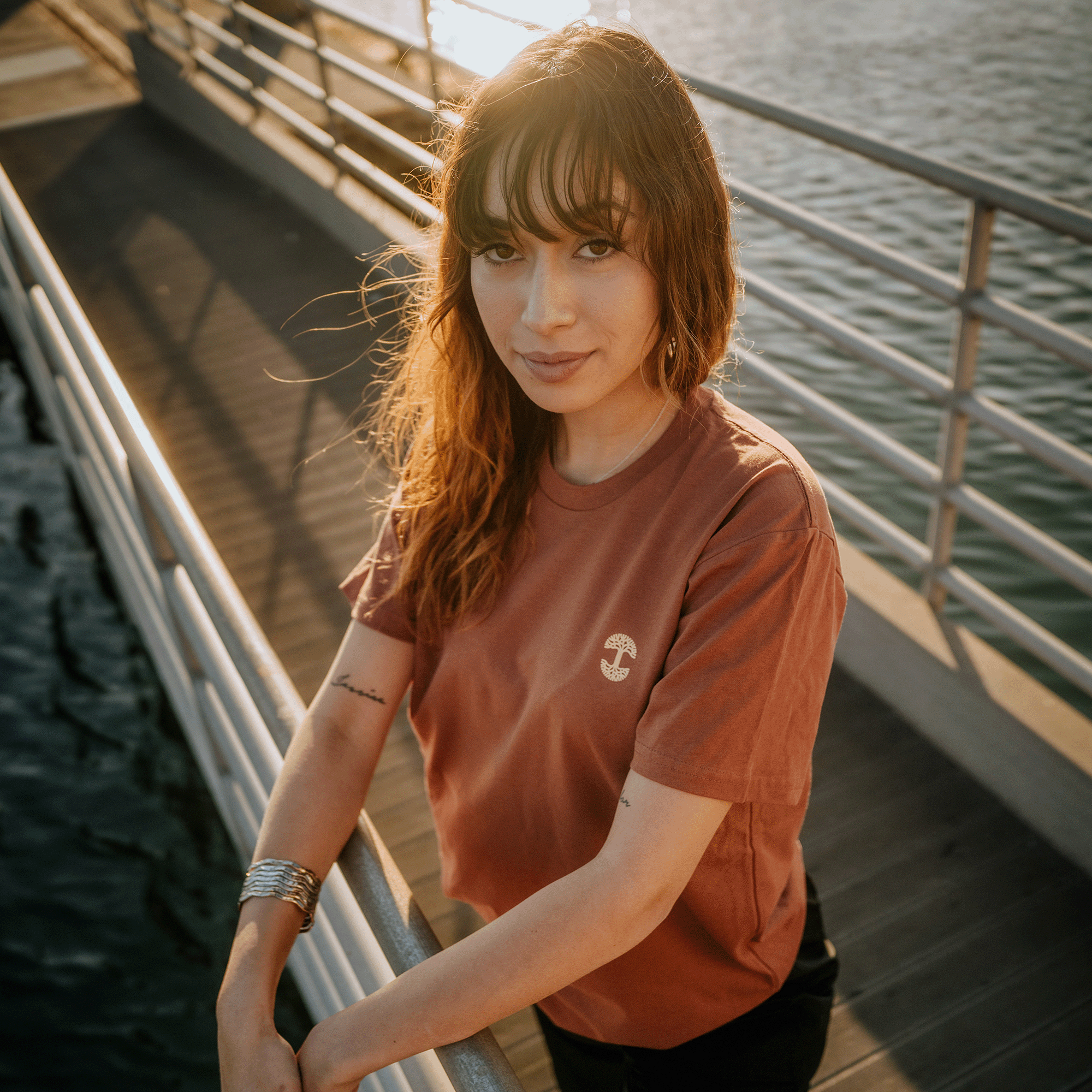 A female model with long brown hair stands on a bridge during sunset, wearing the Peyo Tee from Oaklandish – a rust-colored classic fit t-shirt made from 100% cotton, featuring a small mushroom design. Her forearm tattoos and silver bangle bracelet add to the scene's charm, while the warm sunlight and tranquil river create a peaceful ambiance.