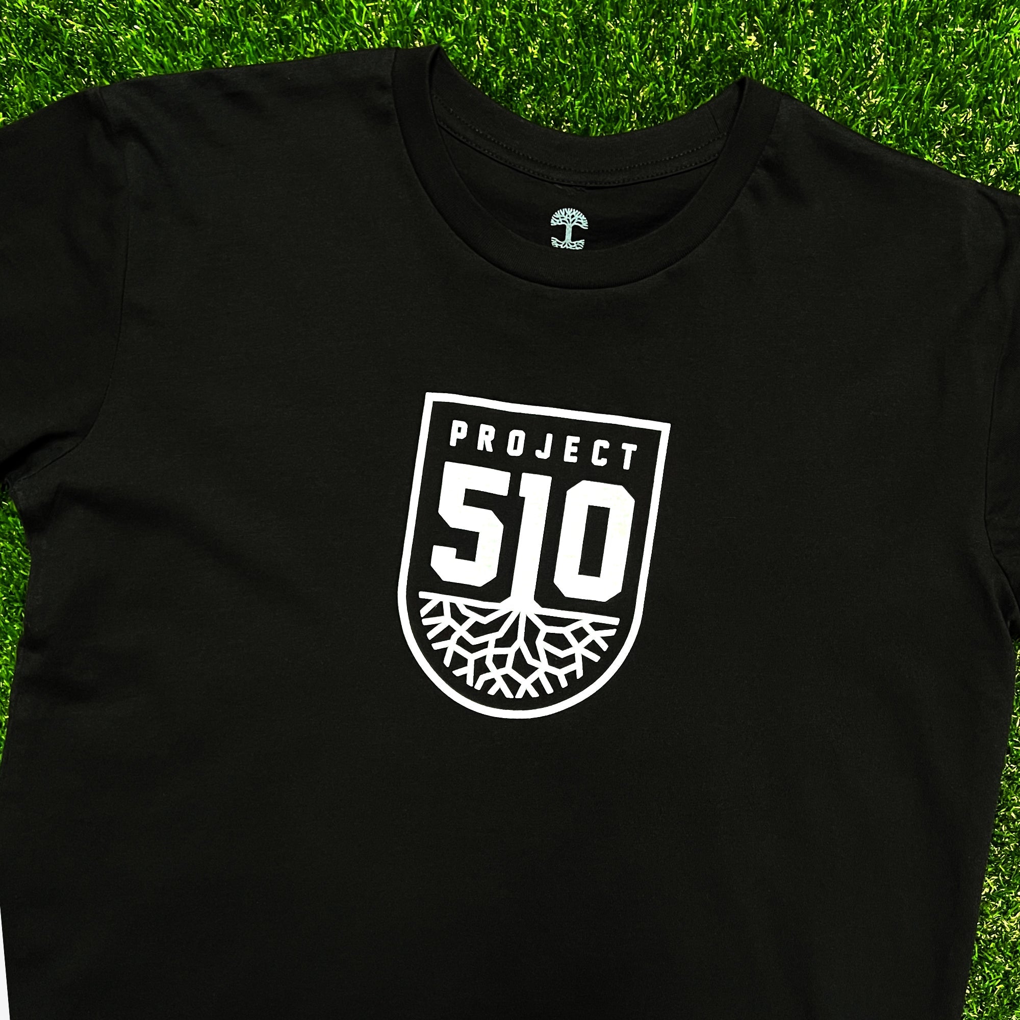 The Project 510 Club Tee by Oakland Roots SC is a black t-shirt that showcases "Project 51O" in bold white letters within a shield-shaped emblem. Below the text, a white graphic depicting tree roots spreads out inside the emblem, hinting at the essence of Oakland Roots SC. The t-shirt is elegantly displayed on a vibrant green grassy background, evoking the feel of a soccer club field.