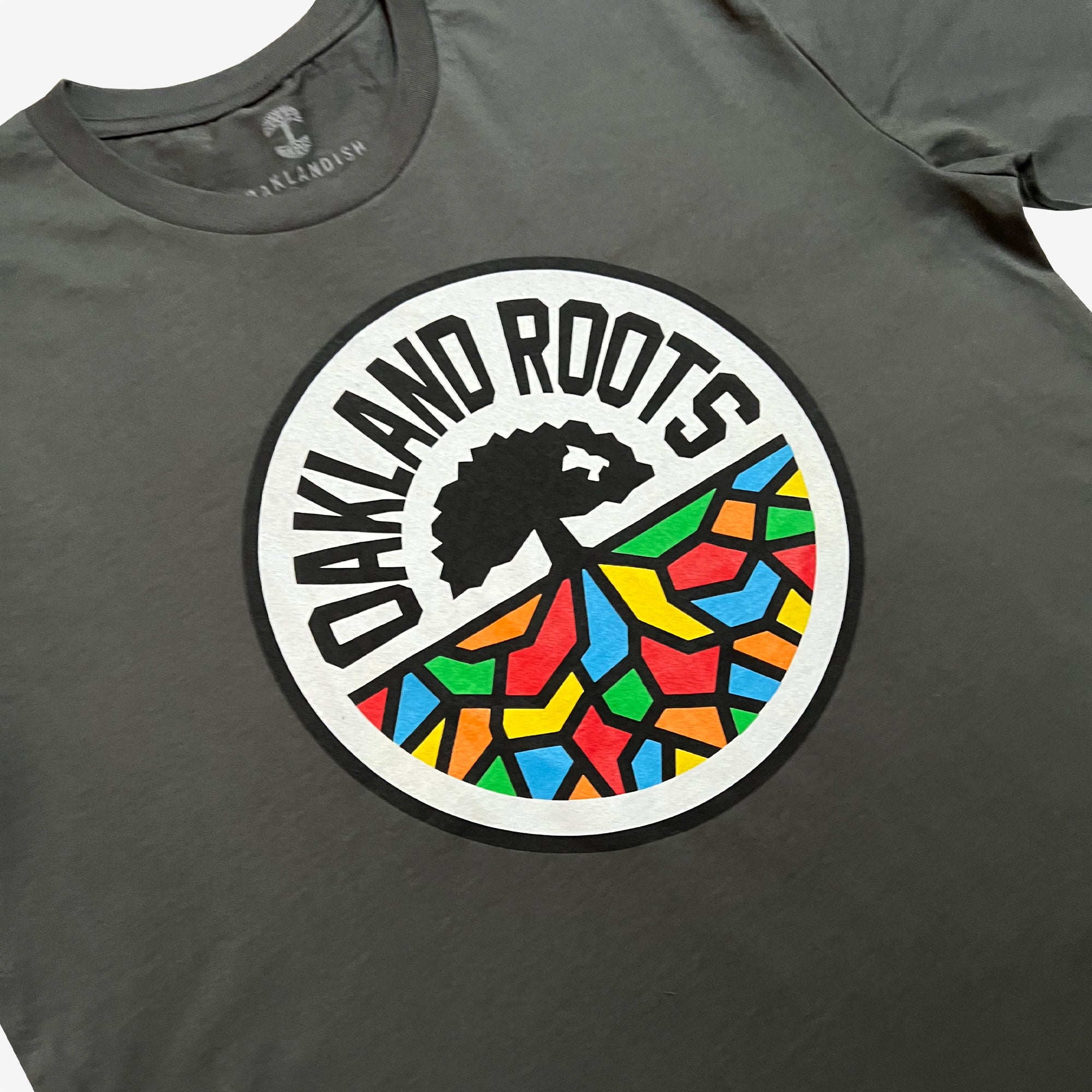 The Oakland Roots SC Classic Tee by Oakland Roots SC is a dark gray T-shirt showcasing a central circular logo. The text "OAKLAND ROOTS" arcs around the top of the circle, while inside, a black tree with spreading roots overlays a vibrant mosaic of multicolored, irregular geometric shapes in red, blue, yellow, green, and orange. The tag reads "OAKLANDISH Sports Club.