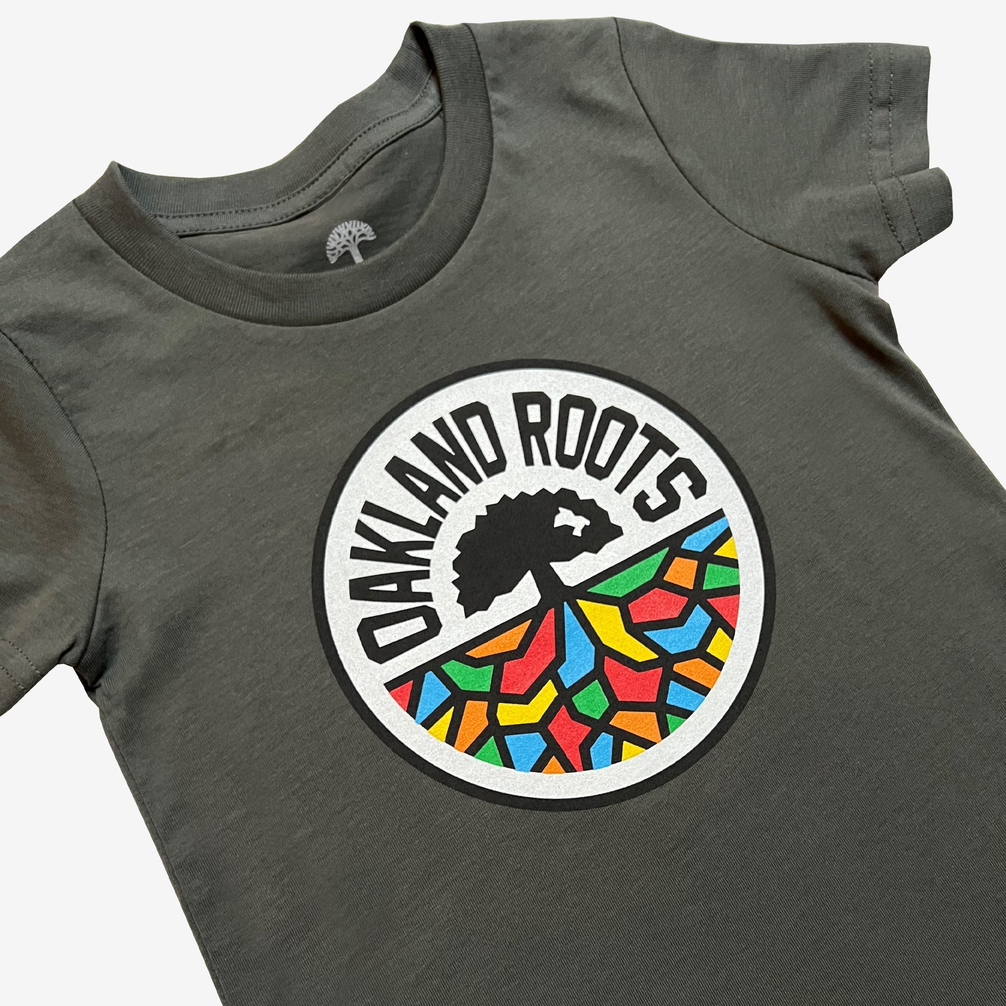 The Toddler Oakland Roots SC Classic Tee by Oakland Roots SC is a dark gray T-shirt adorned with a circular logo at the center. The logo showcases a black tree with spreading roots that transition into a vibrant mosaic pattern, representing the Oakland Roots Sports Club. The words "OAKLAND ROOTS" are arched above the tree within the circle, and an icon of a tree is printed inside the neckline.