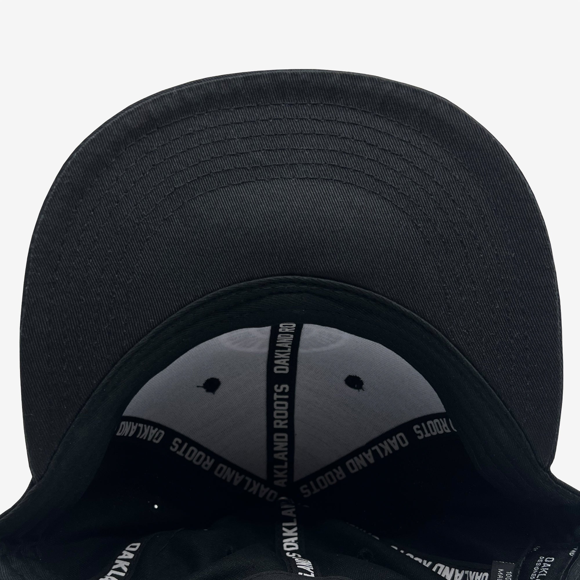 A close-up view of the underside of the Oakland Roots SC Structured Snapback. The inside features white stitching and gray grommets. The inner sweatband is black with repeated white text reading "OAKLAND ROOTS," reflecting its deep connection to Oaklandish culture. The bill of the cap faces towards the top of the image.