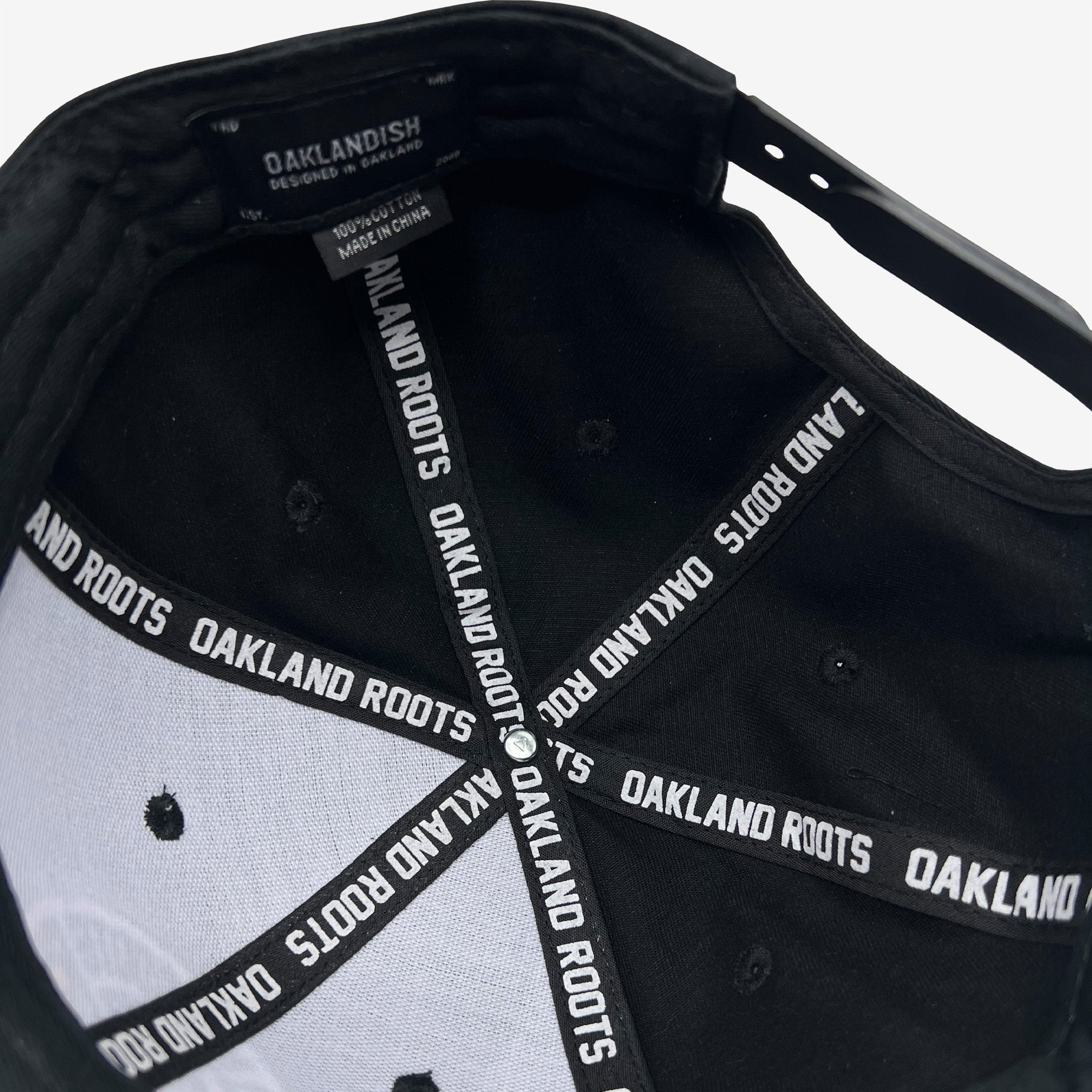 The image displays the interior of an Oakland Roots SC Structured Snapback from the brand Oakland Roots SC. The inside features black fabric tape repeatedly imprinted with "OAKLAND ROOTS" in white, honoring the local sports club. The label inside provides washing instructions and manufacturing details. The snapback closure is also visible.