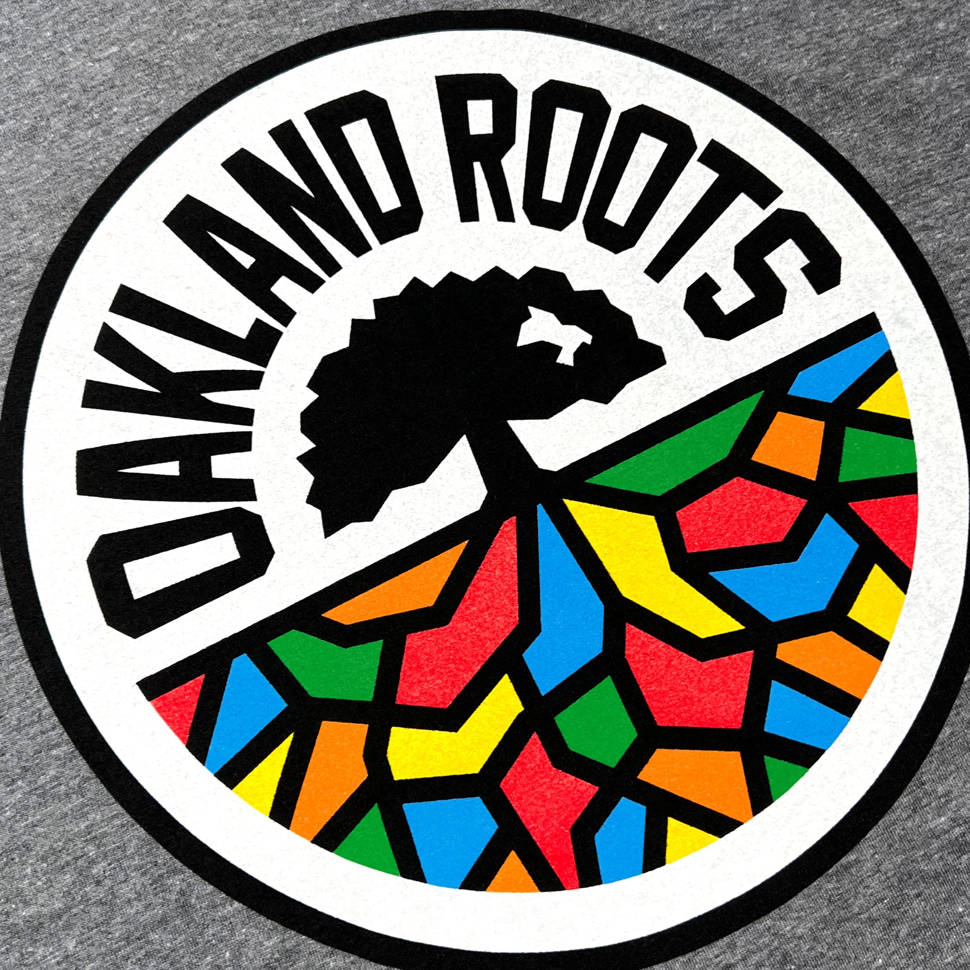 A circular logo with a black outline featuring the text "OAKLAND ROOTS" in uppercase black letters along the top edge. Inside, there's a black tree silhouette rooted in a vibrant mosaic pattern consisting of red, blue, yellow, orange, and green shapes against a gray background, symbolizing Oakland Roots SC's dedication to social good. This design is showcased on the Oakland Roots SC Classic Tee by Oakland Roots SC.