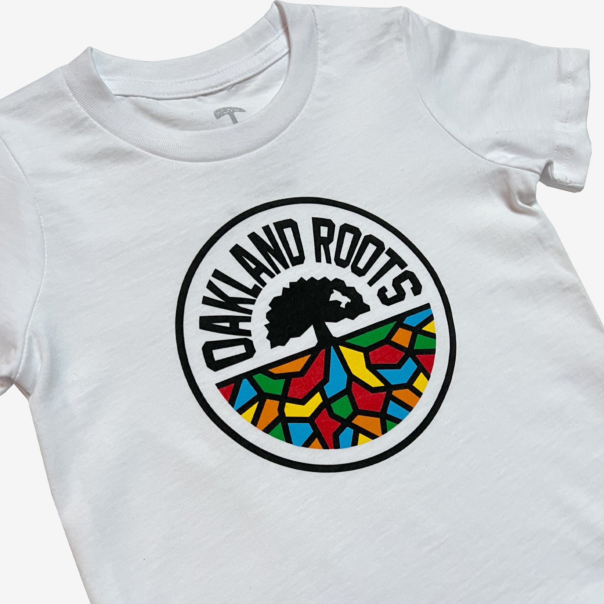 The Toddler Oakland Roots SC Classic Tee displays a circular emblem in the center. The logo, featuring "OAKLAND ROOTS," arches over a silhouetted tree. Below the tree, there is a vibrant mosaic pattern with red, yellow, blue, and green colors that reflects the spirited nature of Oakland Roots SC.