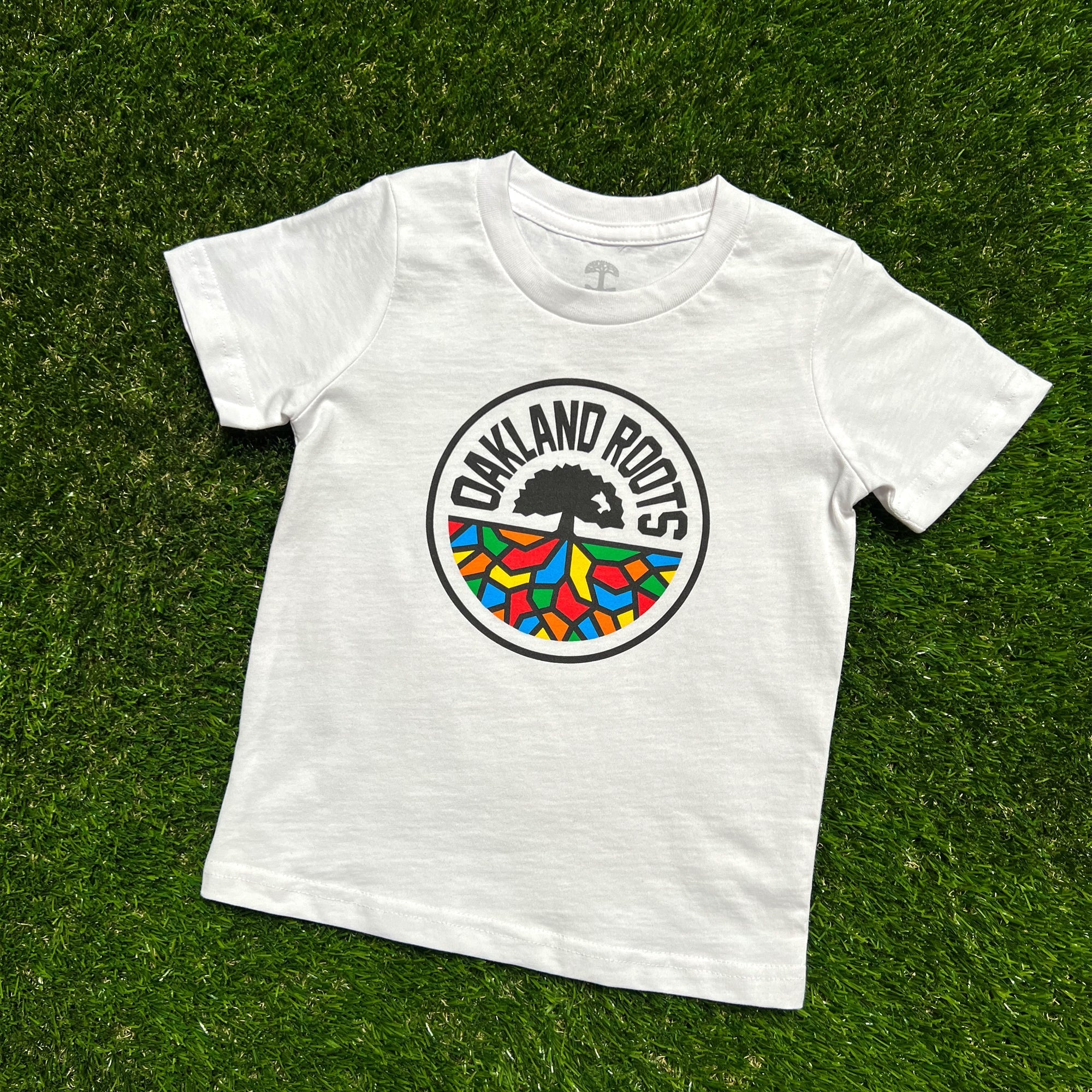 A Toddler Oakland Roots SC Classic Tee featuring the Oakland Roots soccer team logo is displayed on green grass. The circular logo includes "Oakland Roots" text around a black silhouette of a tree, with colorful mosaic-like shapes at the bottom in red, yellow, blue, and green—a perfect blend of Oaklandish flair and Sports Club pride.