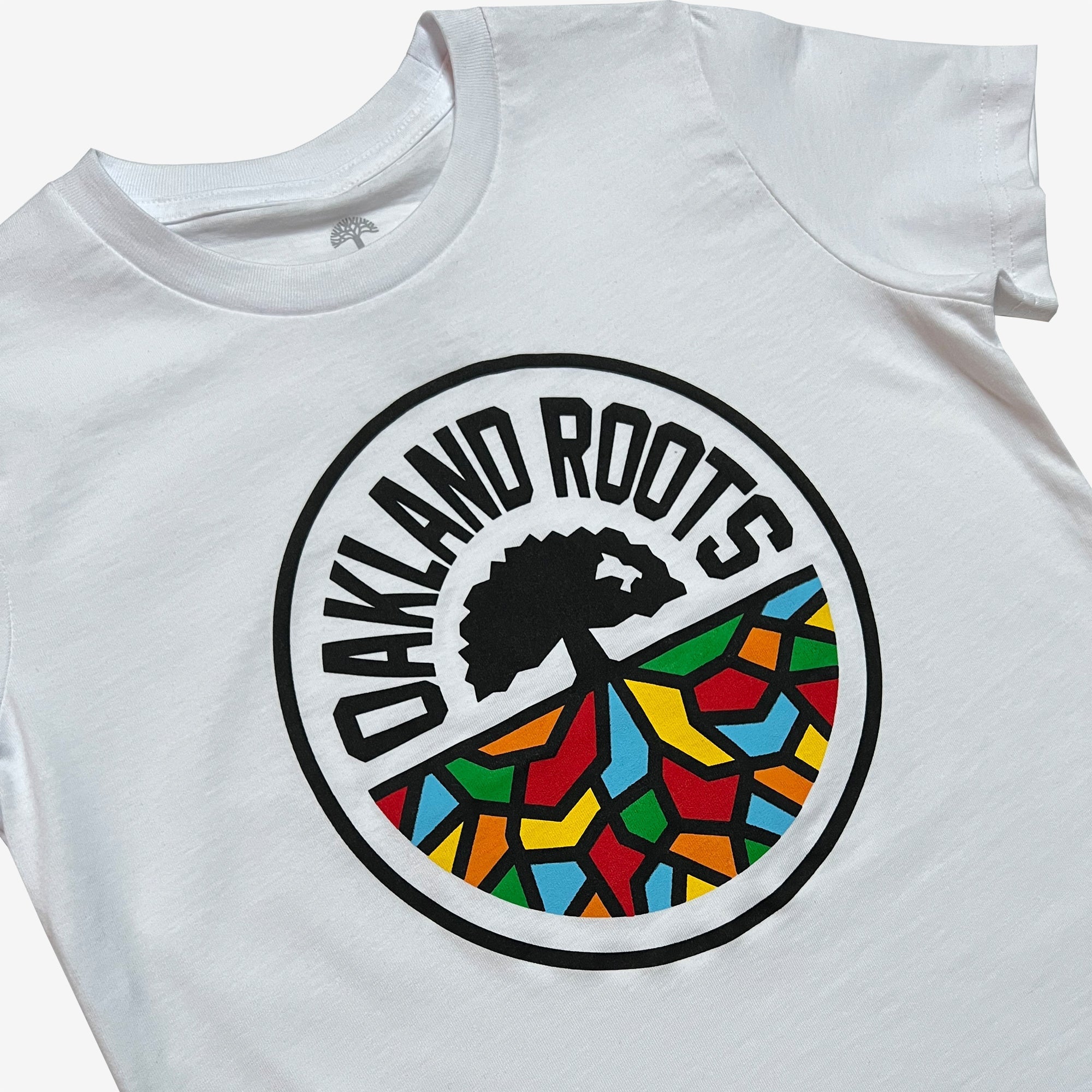 The Youth Oakland Roots SC Classic Tee by Oakland Roots SC is a white t-shirt featuring a circular emblem on the front. The emblem showcases a black tree silhouette with branching roots over a multicolored mosaic pattern, symbolizing social good. The text "OAKLAND ROOTS," representing the Oakland sports club, is printed in all caps around the top of the circle.