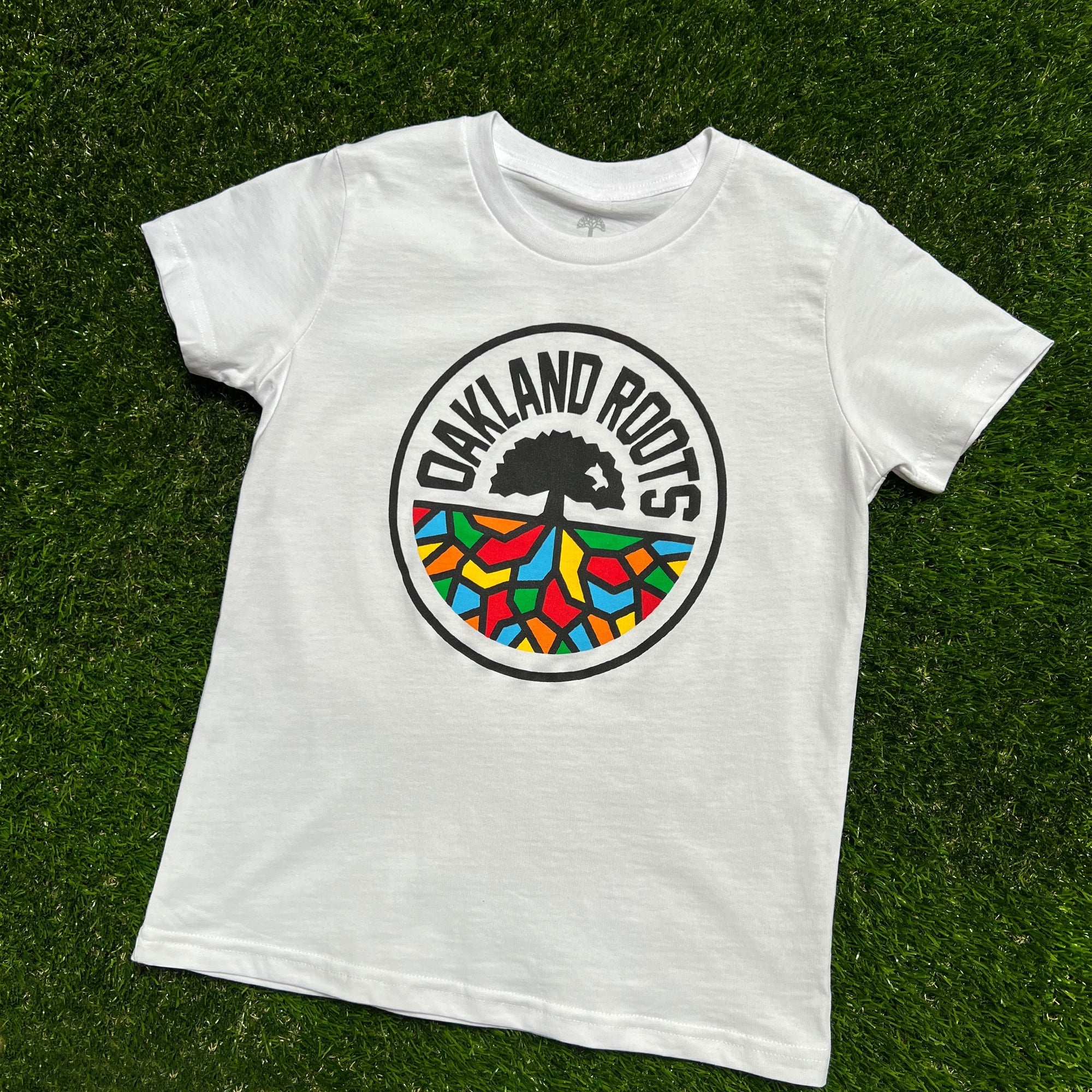 A Youth Oakland Roots SC Classic Tee is displayed lying flat on a green grass background. The T-shirt features a black-and-white circular logo in the center with the words "OAKLAND ROOTS" above a black tree. Beneath the tree is an area resembling multi-colored stained glass, reflecting the vibrant spirit of the Oakland sports club.