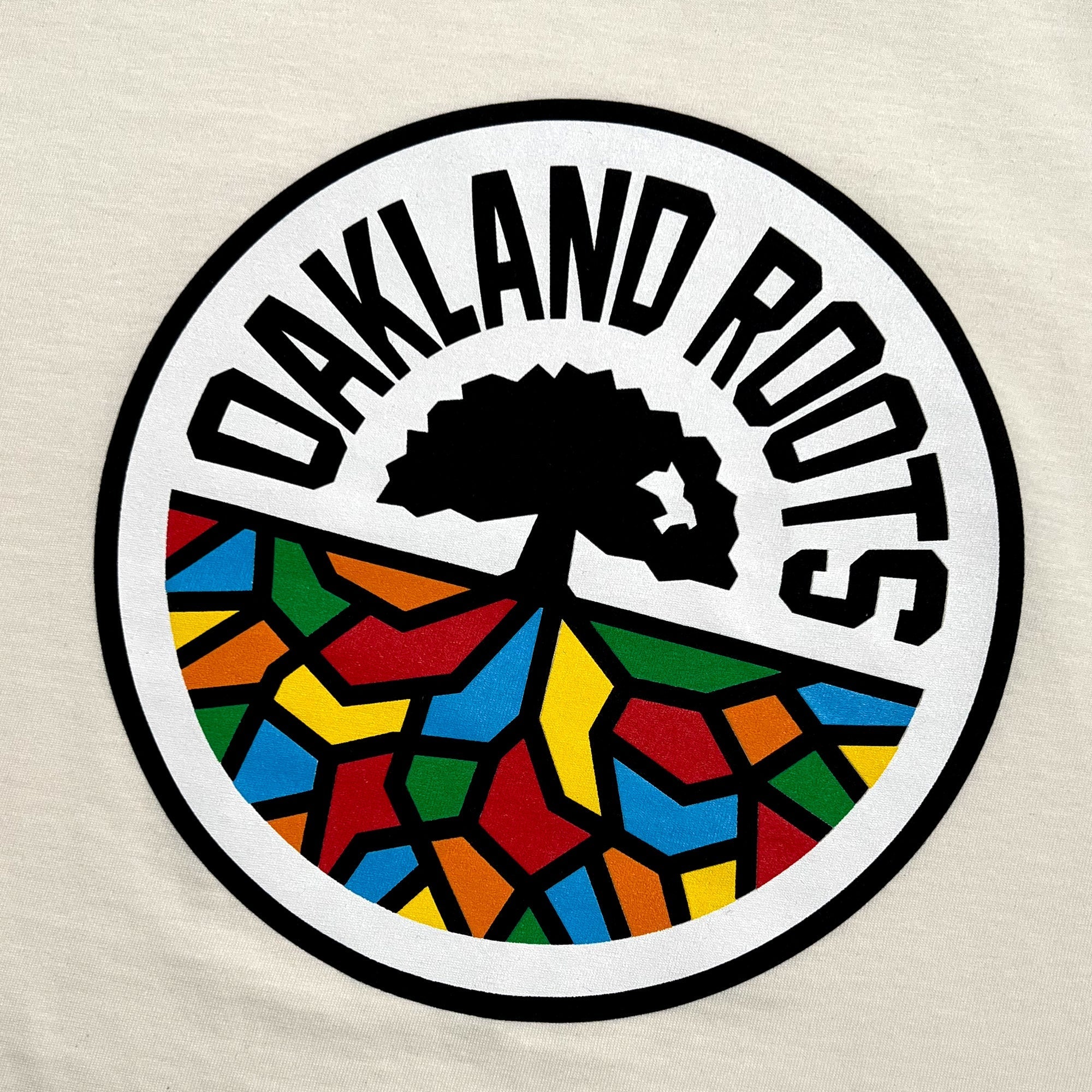 The Women's Oakland Roots SC Classic Tee features a circular logo with the bold black text "OAKLAND ROOTS" curving around the top. Inside the circle, there's a black tree with widespread roots depicted, and the bottom half contains a colorful, mosaic-patterned ground in shades of red, yellow, blue, green, and orange. It's perfect for any fan of Oakland sports clubs. This tee represents the Oakland Roots SC brand proudly.