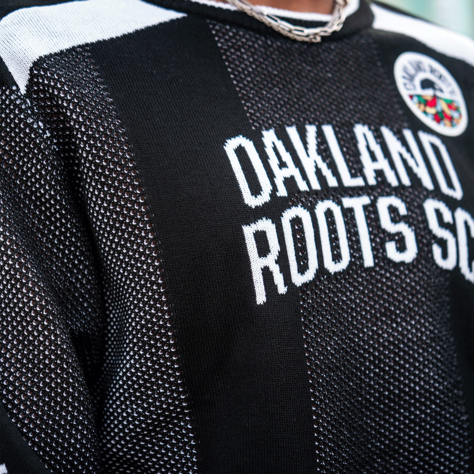 A close-up of the Oakland Roots SC Clubhouse Sweater shows black and white textured fabric, "OAKLAND ROOTS SC" in bold white on the front, a colorful round logo patch on the upper right chest, and a silver chain necklace at the neck.