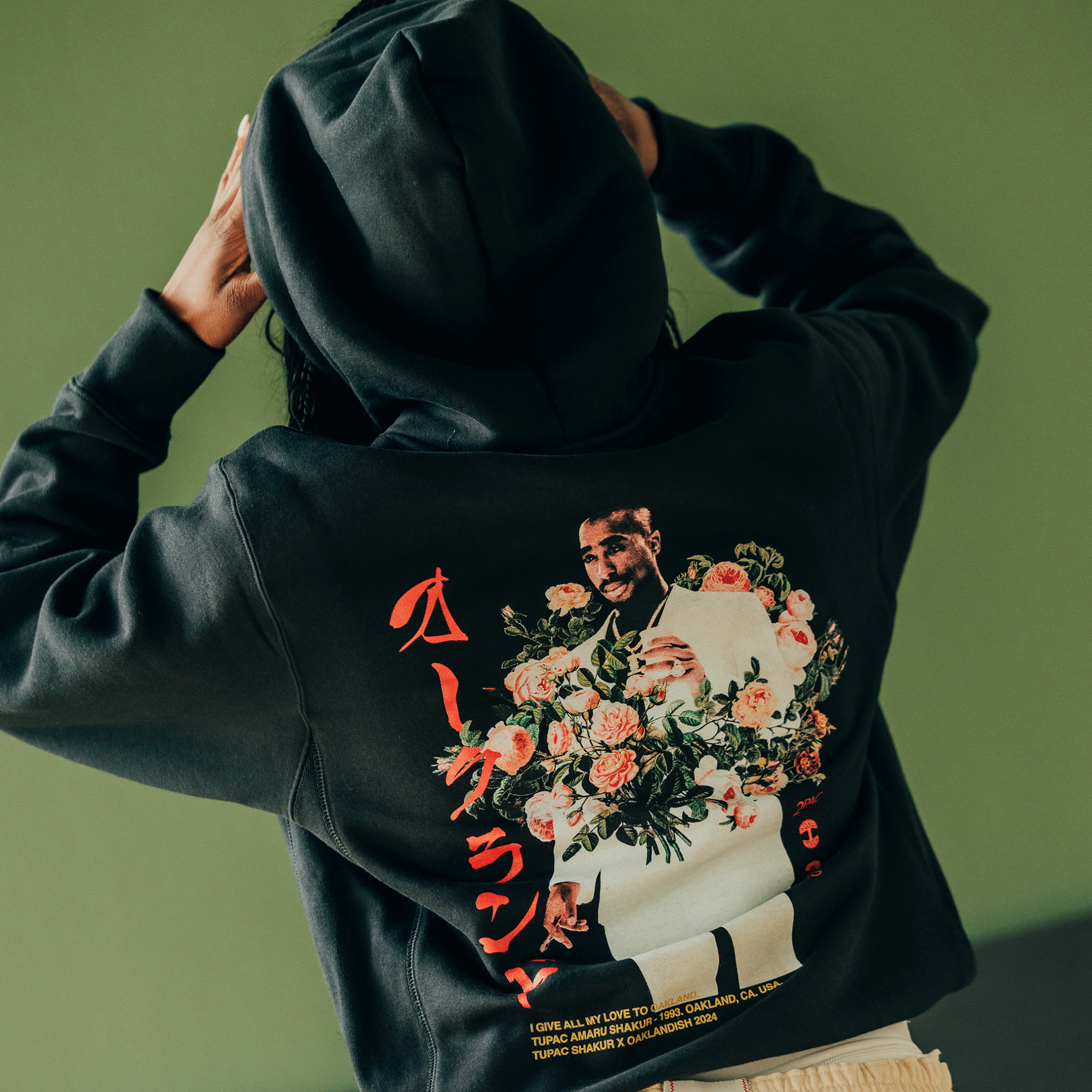 Wearing the Roses Hoodie from Oaklandish, this piece honors Tupac's legacy with a back illustration featuring a smiling man encircled by flowers. The design is accentuated by red and white text in a stylized font, set against a plain green backdrop as their hands rest on their head.